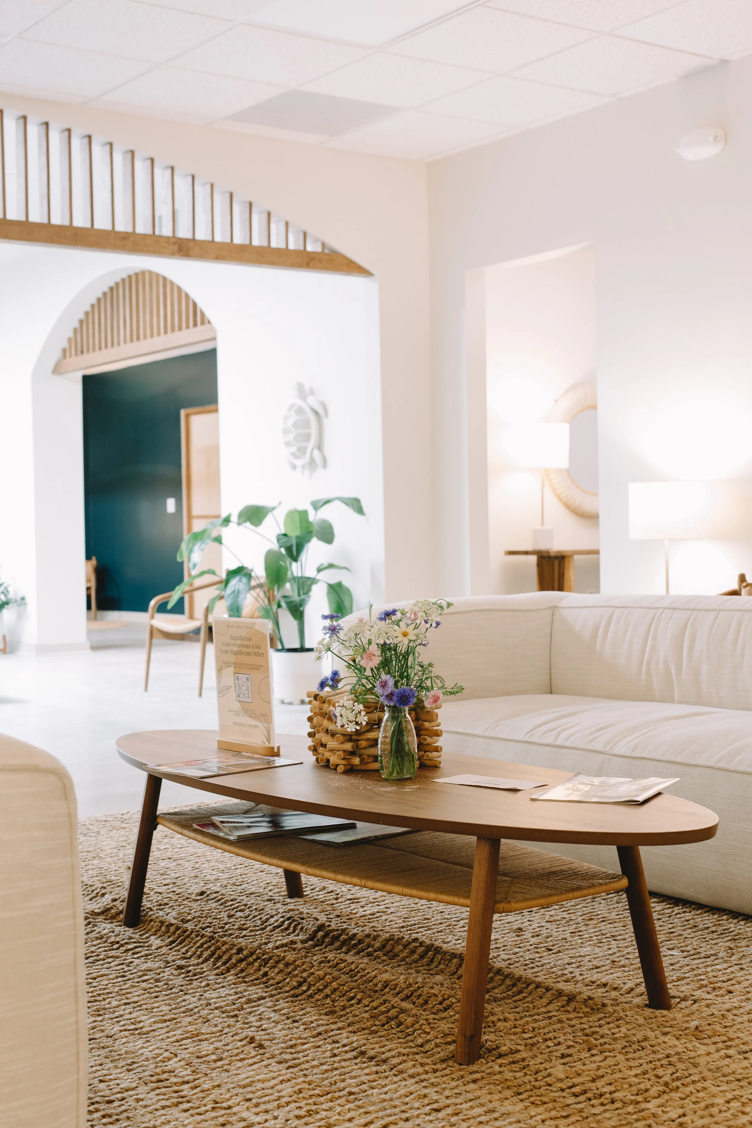 A cozy, well-lit living room with beige and cream tones, decorated with a wooden coffee table, a cream sofa, a large textured area rug, a bouquet of flowers, and decorative wall art. Behind the sofa, a wooden side table holds a lamp. There are plants and additional seating visible in the background, with an archway leading to another room.