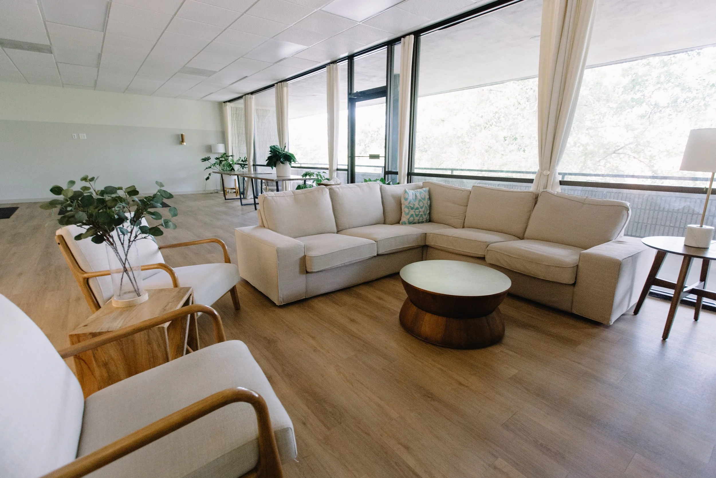 Living room with a beige sectional sofa, wooden coffee table, white armchair with wooden frame, green potted plants, large windows with curtains, and light wood flooring.