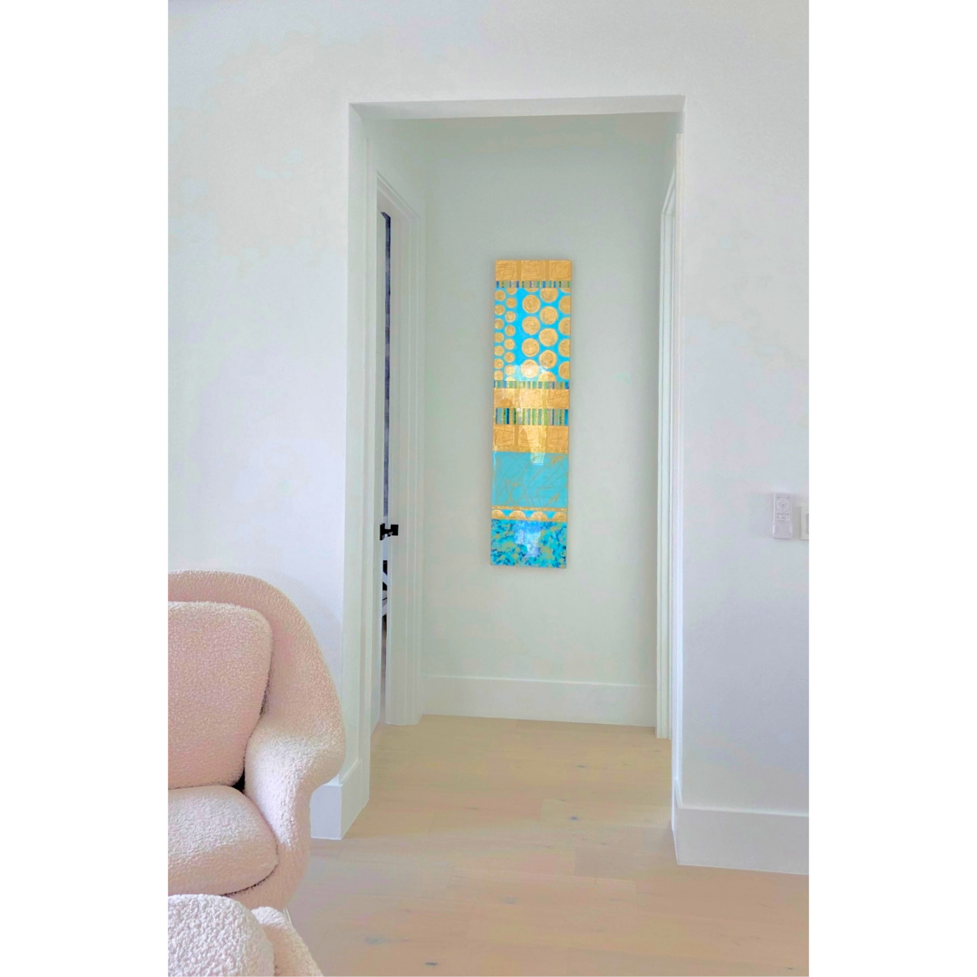 Interior of a room with white walls, a pink upholstered armchair, and a wall hanging with colorful abstract patterns in a narrow vertical frame.