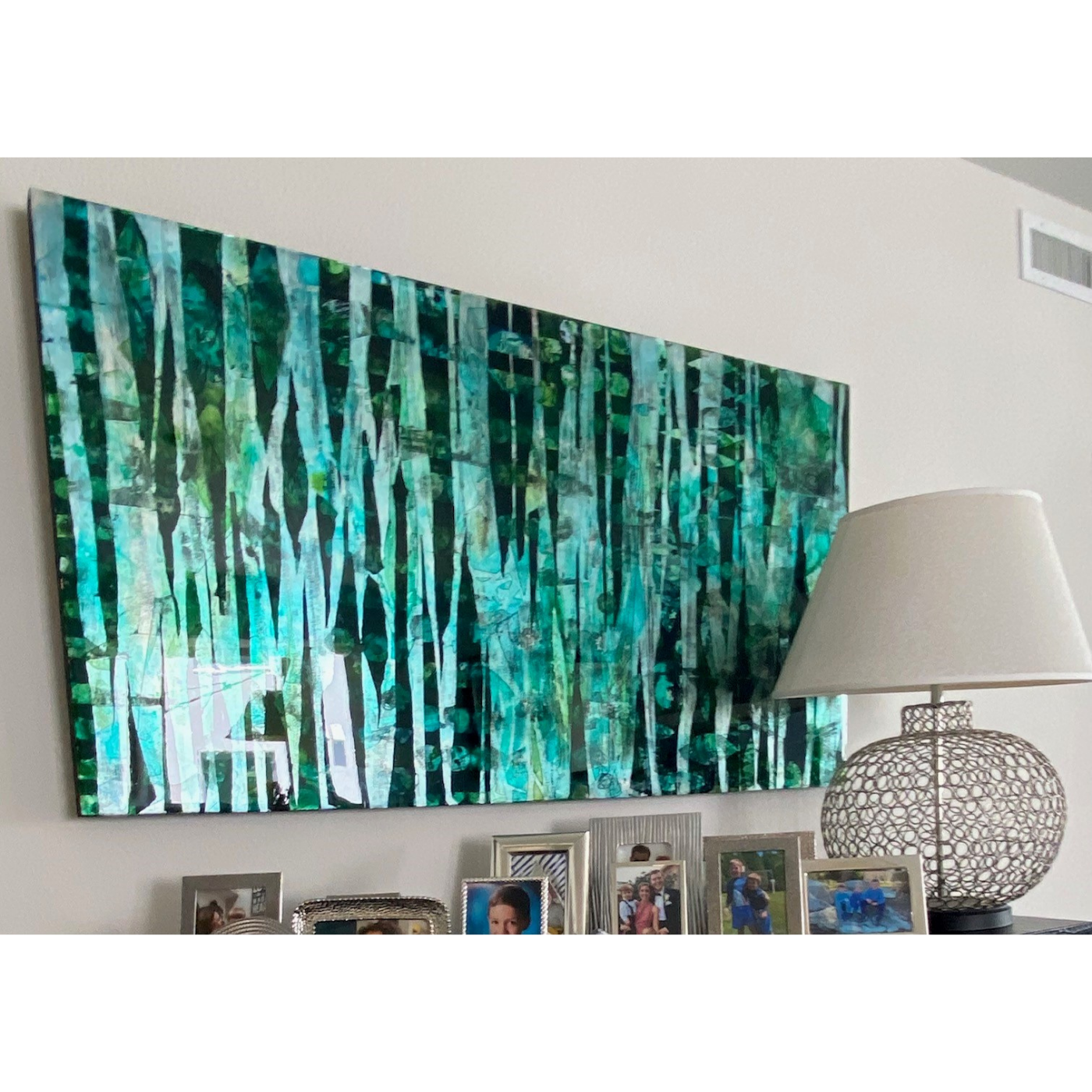 A framed abstract painting of a forest scene with green and blue hues, hanging on a white wall above a sideboard with family photographs and a lampshade.