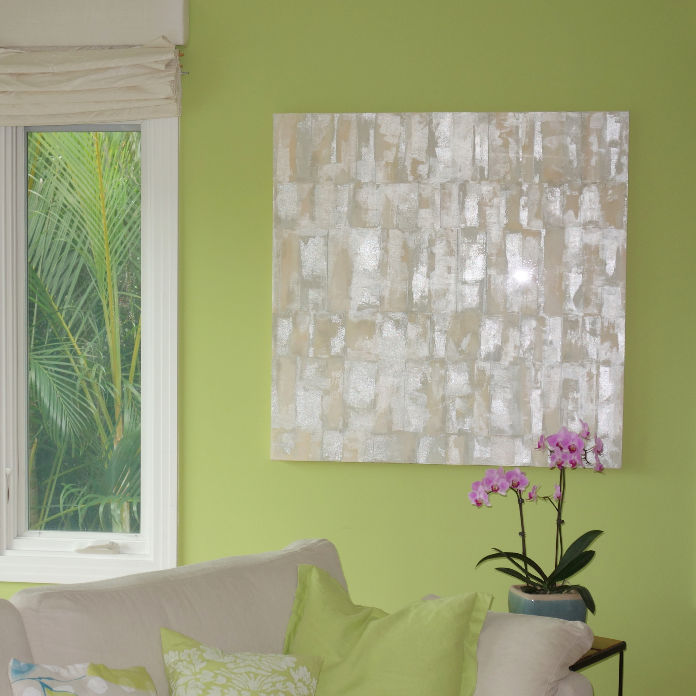 A living room with a lime green wall, a white couch with green and patterned pillows, a window with a view of green foliage, a textured light gray and white wall art, and a potted pink orchid on a small table.