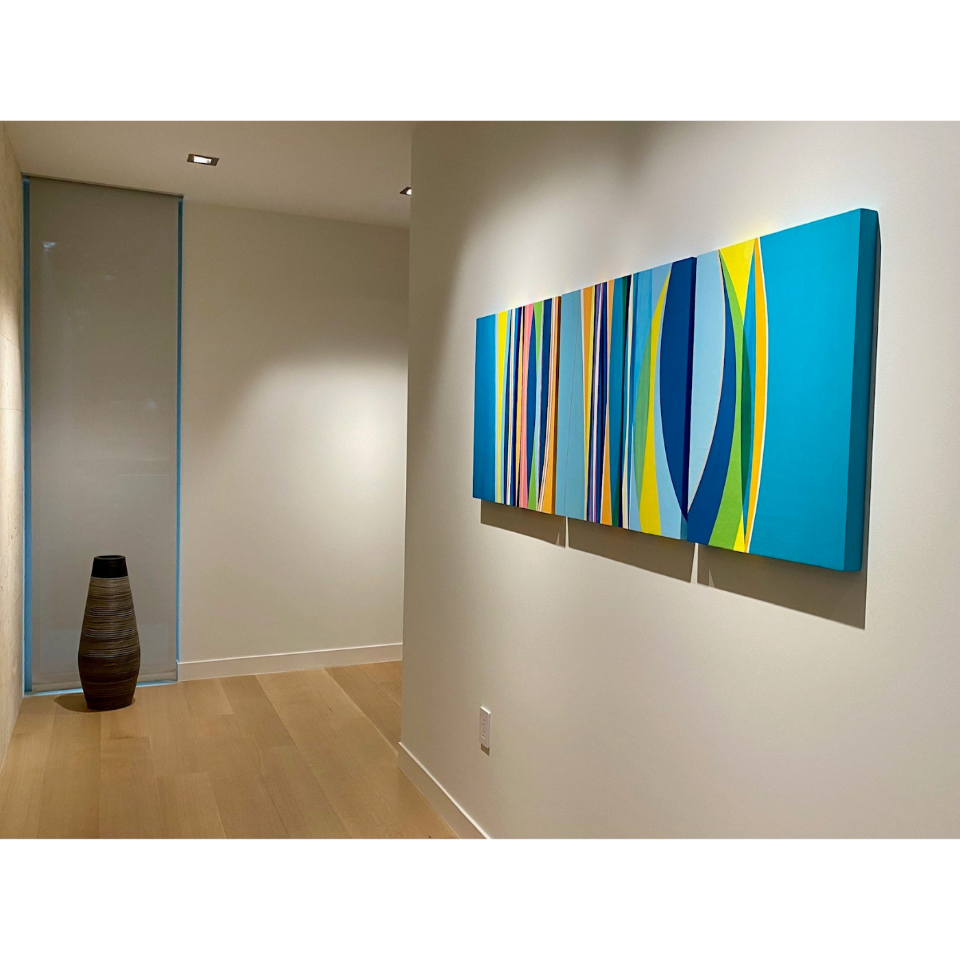 Modern interior with a colorful abstract painting on white wall, a decorative vase on wooden floor, and a glass partition with blue trim.