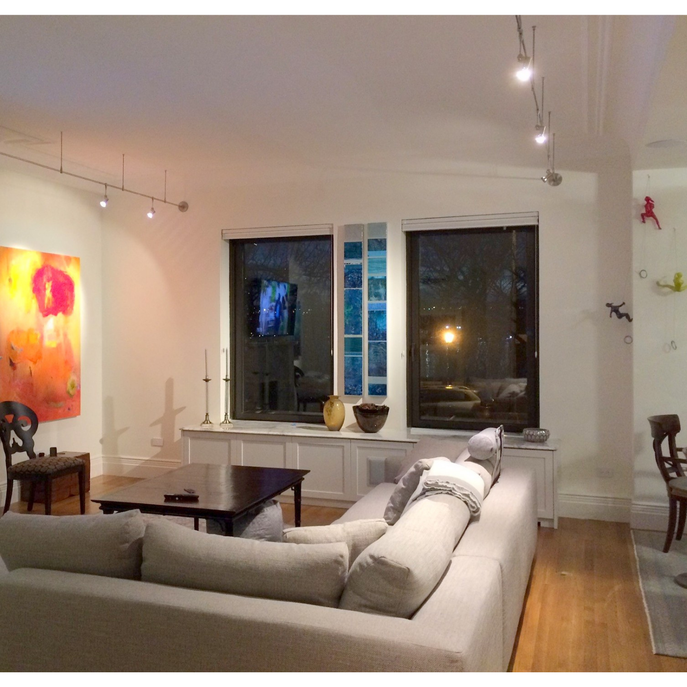 Living room with white sofa, wooden coffee table, artwork on the wall, and large windows at night. There are decorative items on the windowsill and colorful wall sculptures near the corner.
