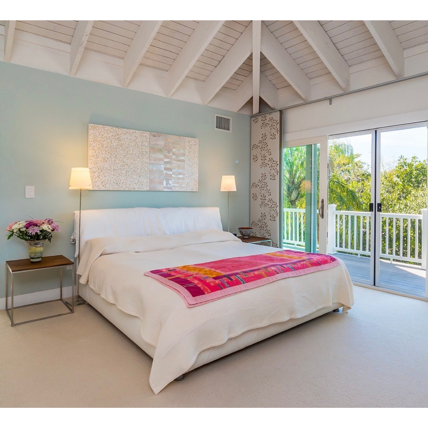 A bedroom with a white bed, pink and orange quilt, two bedside lamps, a nightstand with flowers, a large abstract wall art, sliding glass door opening to a balcony with green trees outside, and a high vaulted ceiling with wooden beams.