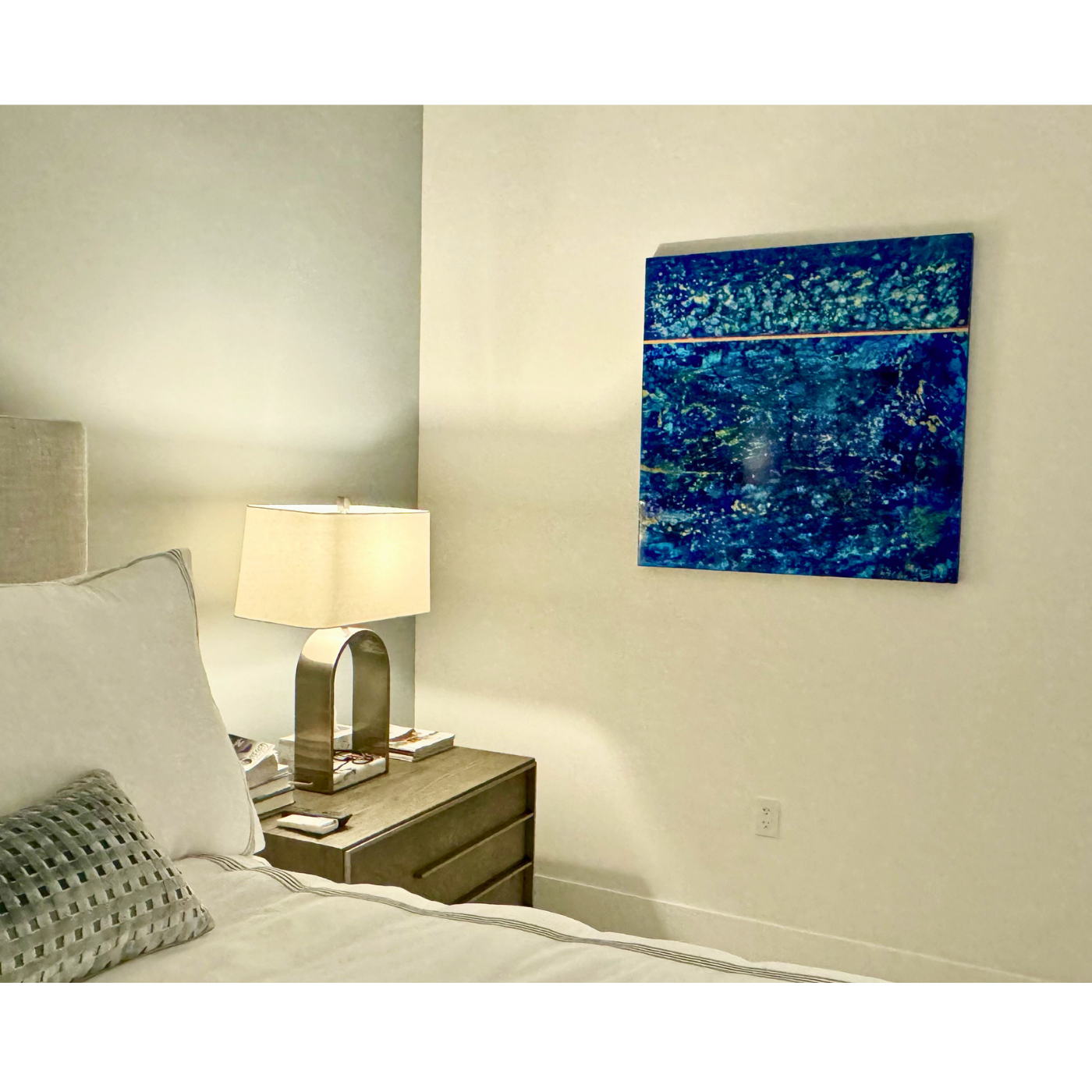 A bedroom with a beige bed, a matching beige pillow with a checkered pattern, a wooden nightstand with a modern silver desk lamp, and a blue abstract painting on the wall.