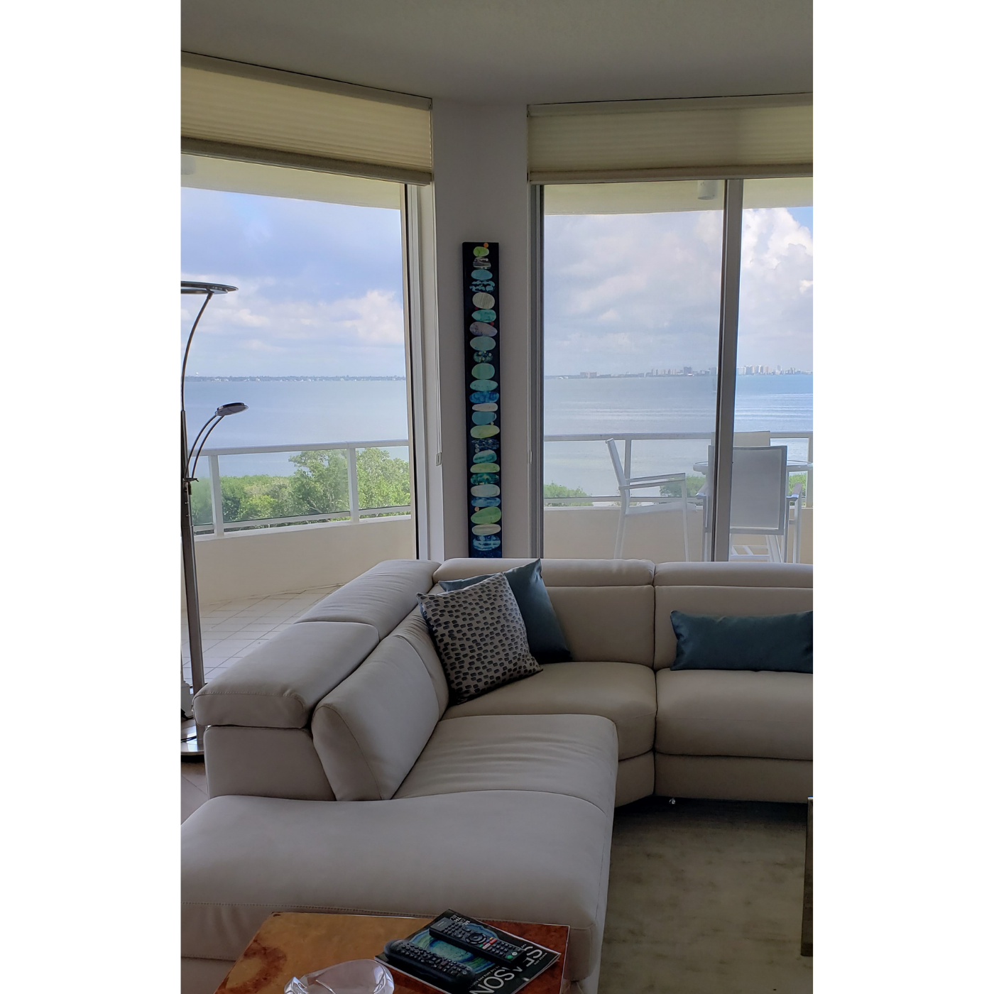 Living room with a white sectional sofa, decorative pillows, large windows with a view of water, a balcony with outdoor chairs, a tall vertical decorative piece, and a lamp.