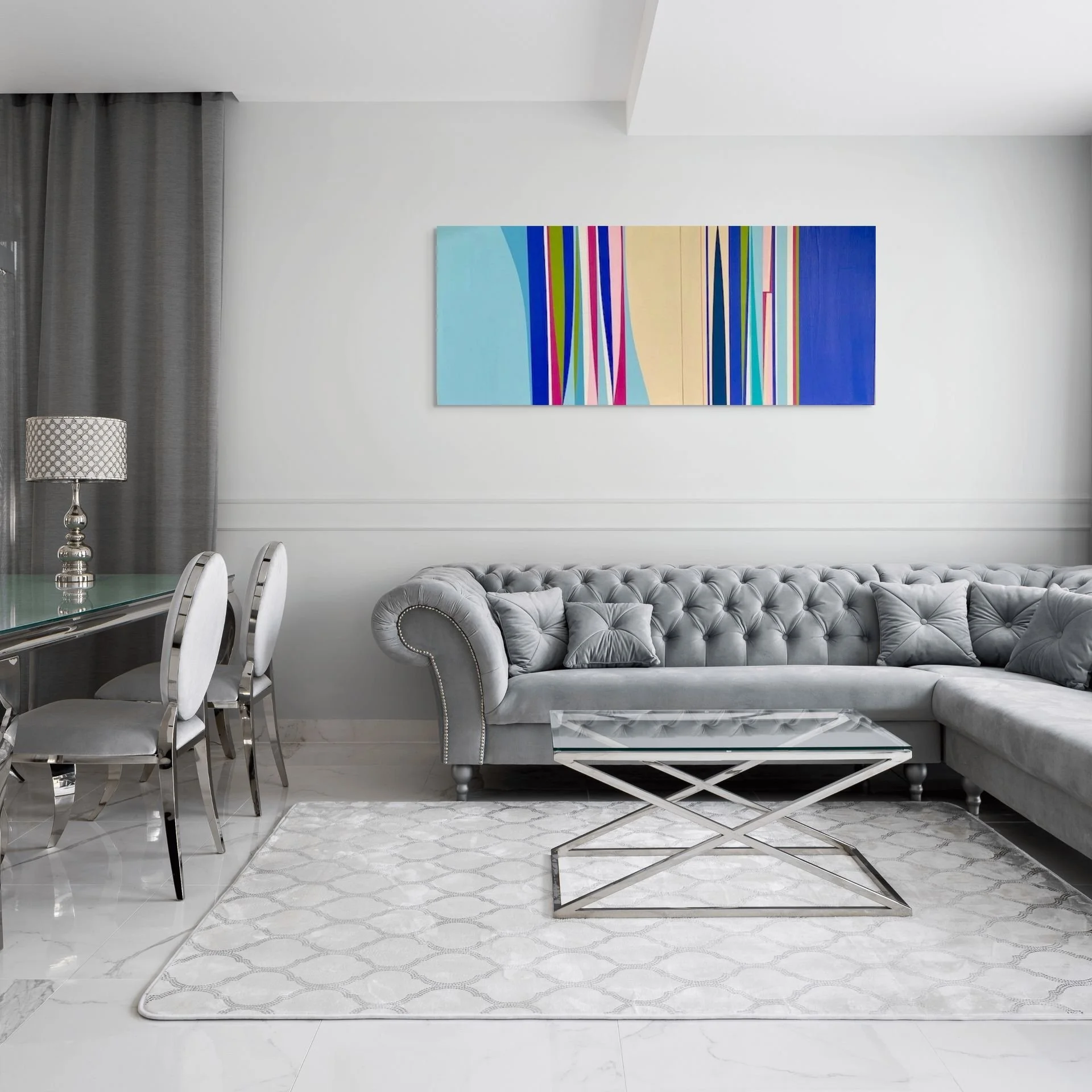 Modern living room with gray tufted sectional sofa, glass coffee table, abstract colorful painting on white wall, gray curtains, and a silver lamp on a glass dining table with white chairs.