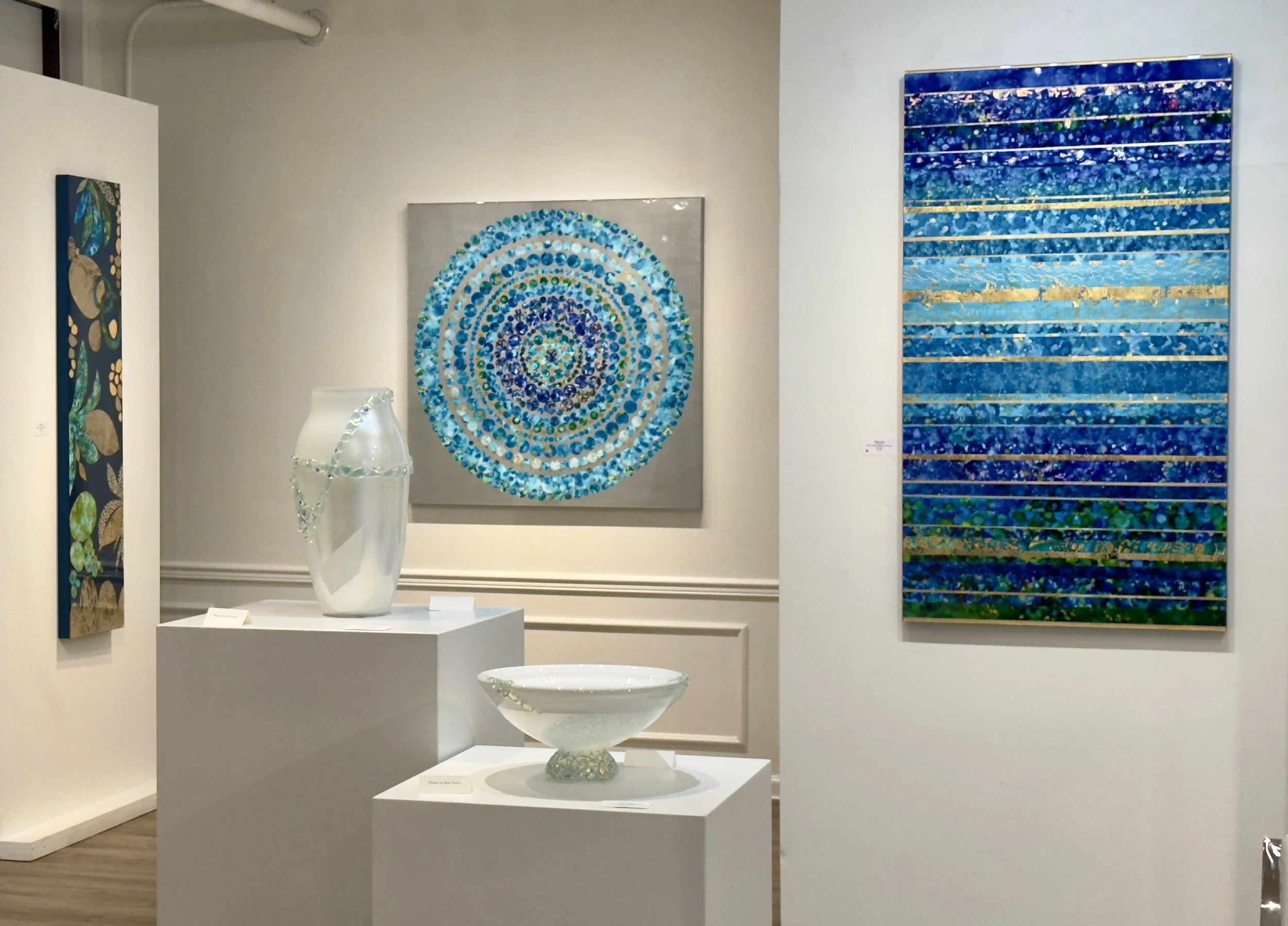 Art gallery featuring abstract paintings, including a circular mosaic composed of blue and green dots on a gray background, another blue mixed-media piece with horizontal stripes, and decorative ceramic sculptures in white on white pedestals.