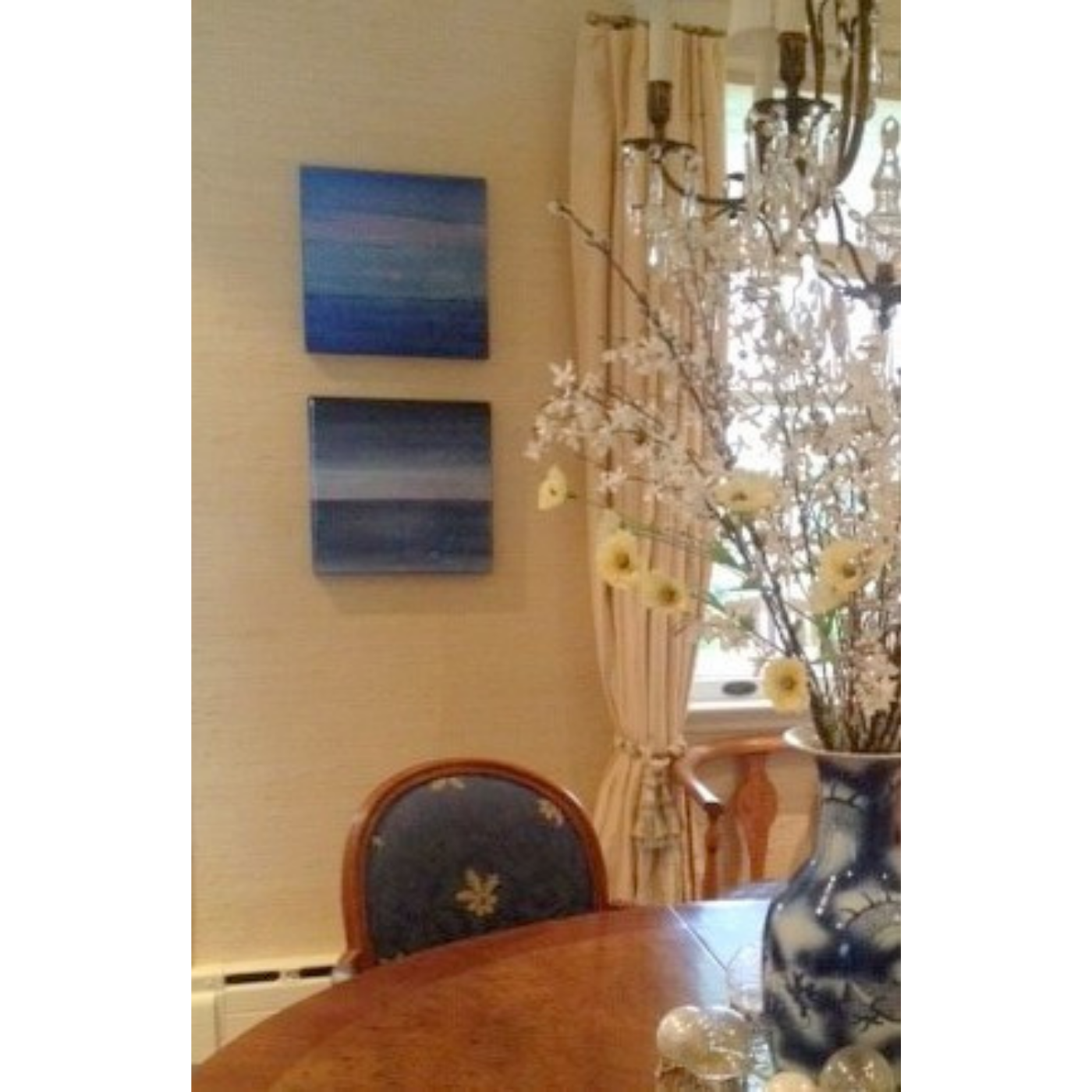 A dining room with a wooden table, a decorative vase with flowers, and chairs. The wall has two abstract paintings and a window with curtains. A chandelier hangs from the ceiling.