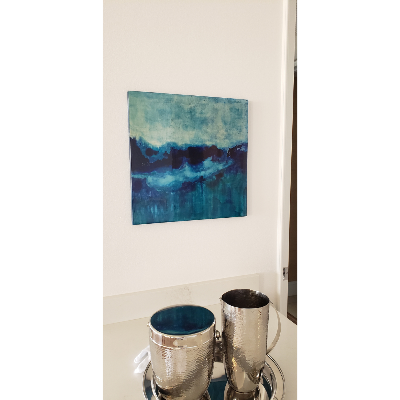 Decorative table with metallic silver cups and tray, in front of a white wall with a blue and green landscape painting.