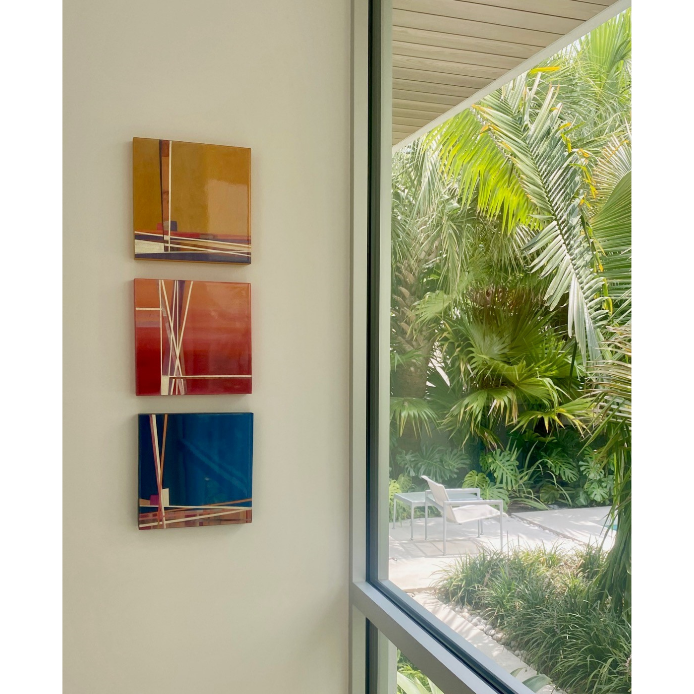 Interior wall with three abstract colorful paintings hung vertically next to a large window showing lush tropical plants outside and a white outdoor lounge chair.