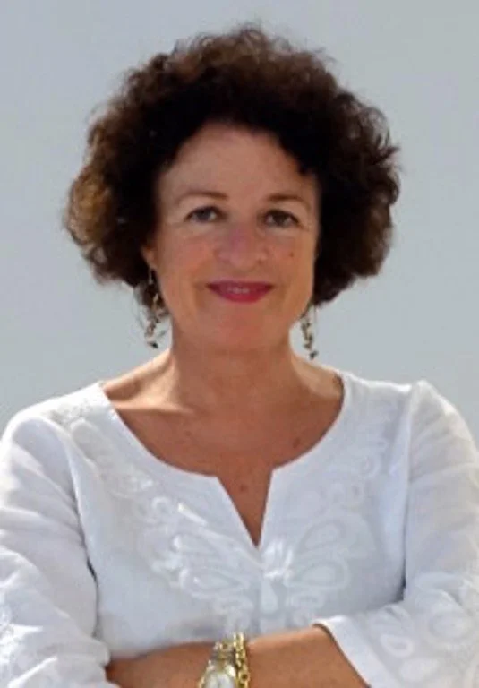A woman with curly brown hair, wearing a white top, smiling, with her arms crossed.
