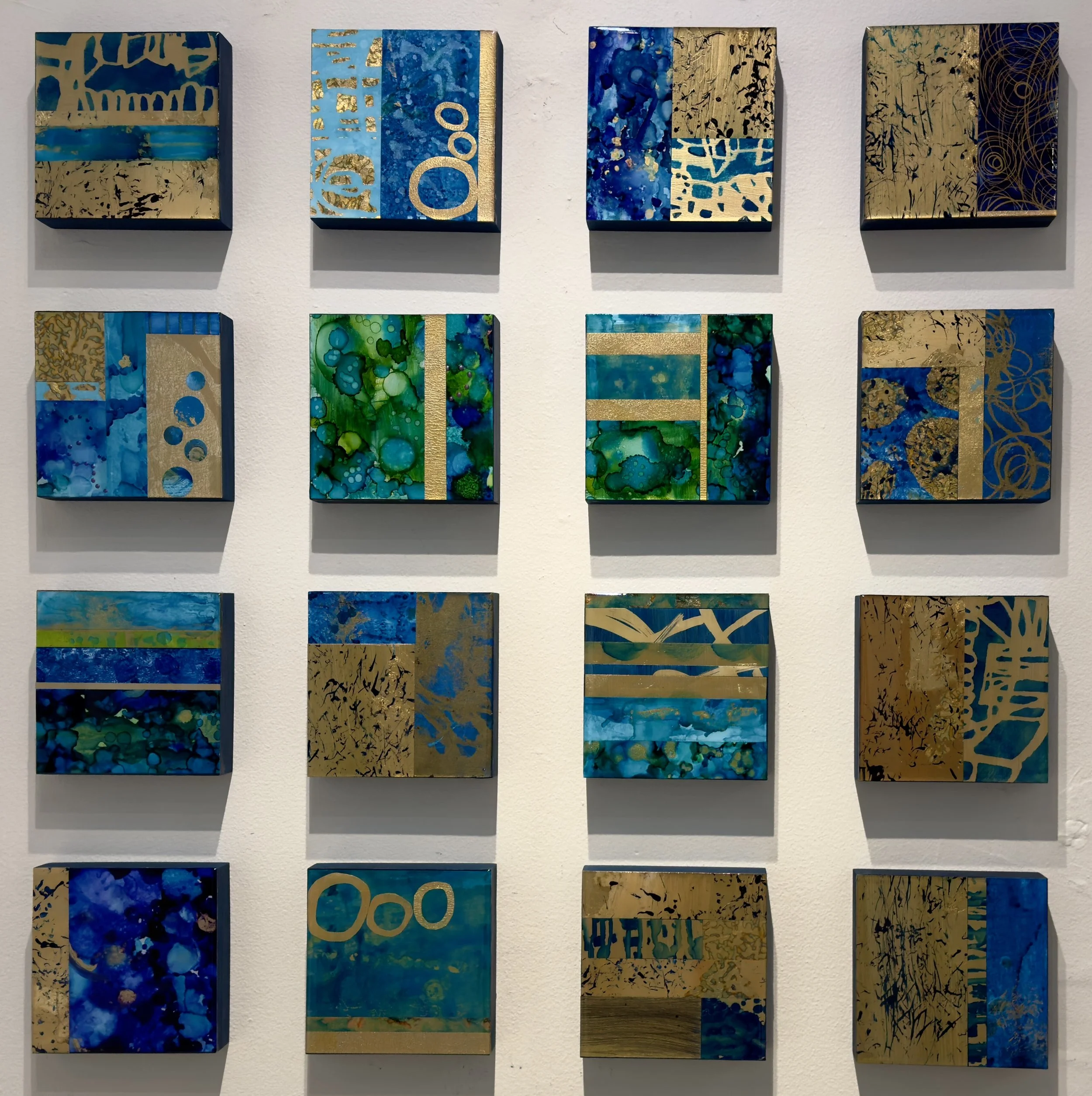 A wall display of 15 small square abstract paintings featuring blue, green, and gold colors with various patterns and textures.