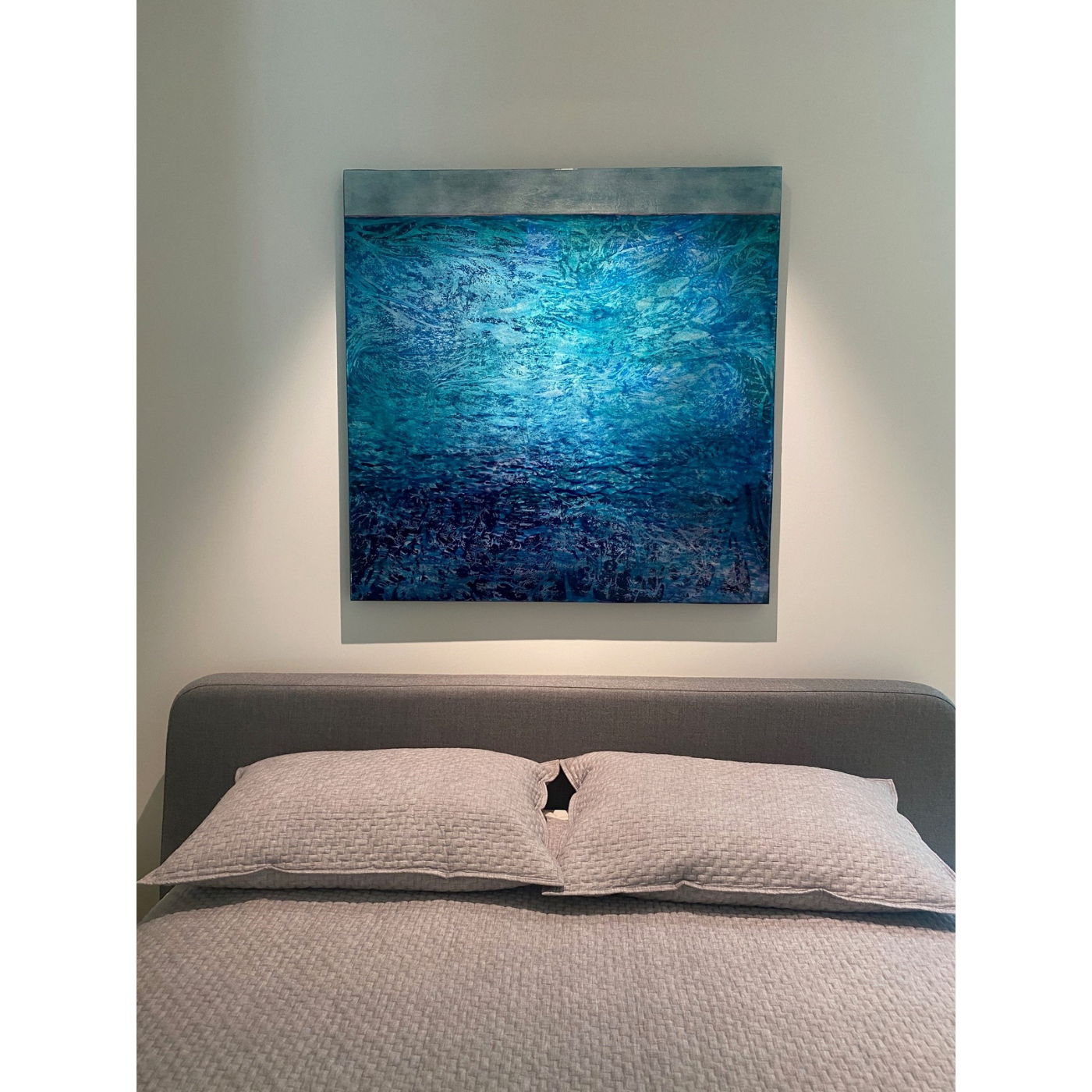 A bedroom with a gray upholstered bed and beige quilted bedding. Above the bed is a large abstract blue painting on a white wall.