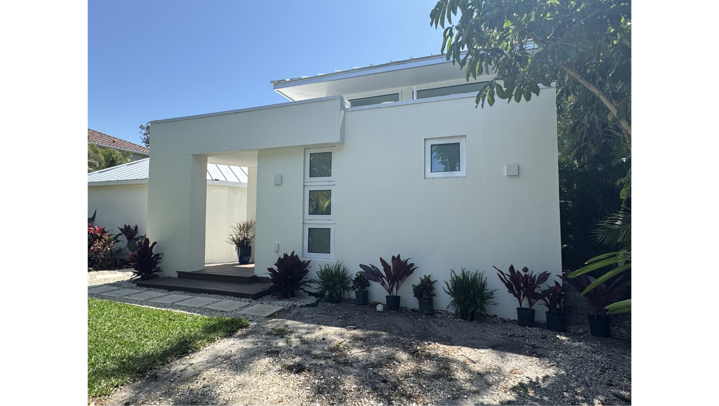 Modern white house with minimalist design, small front steps, potted plants, and surrounding greenery, under a blue sky.