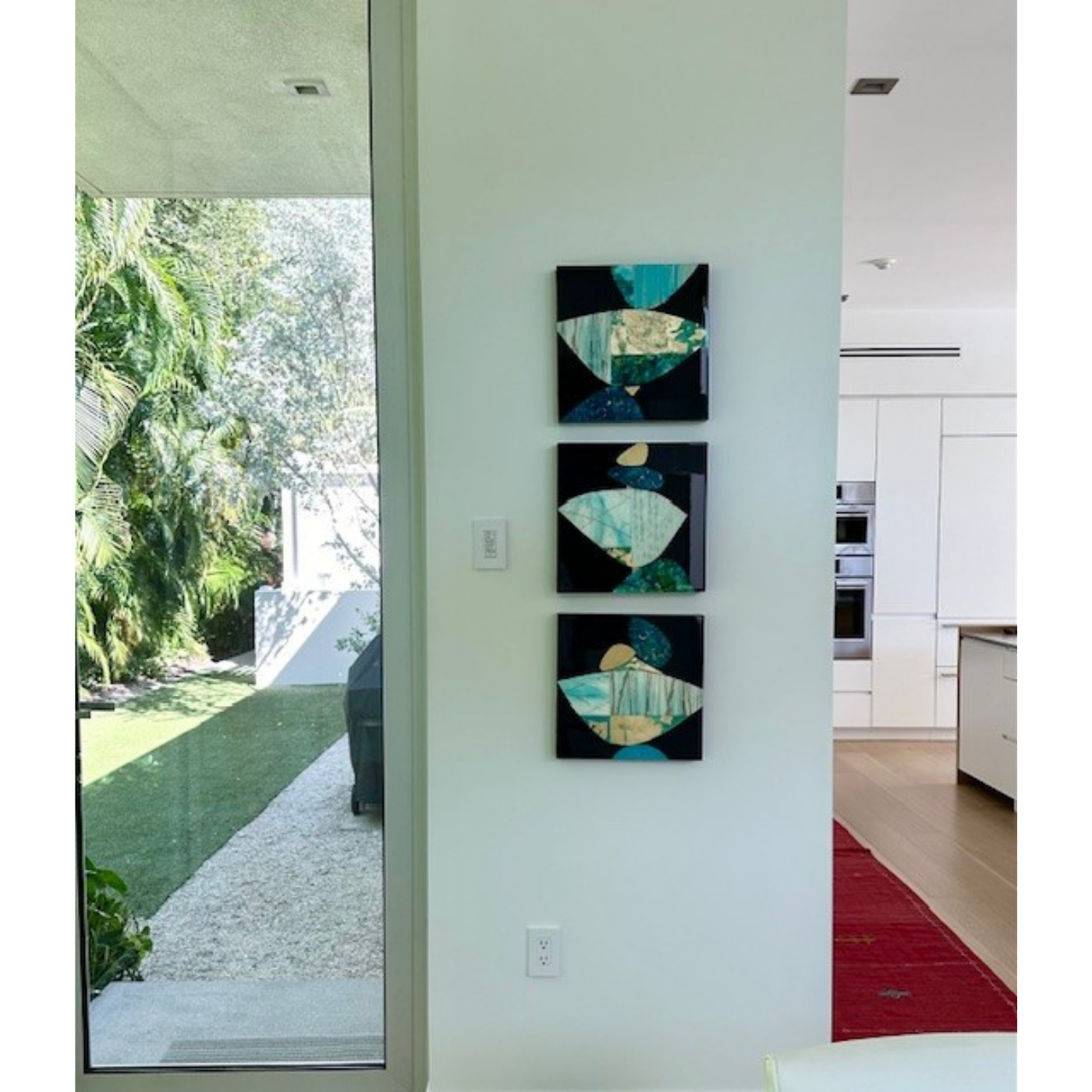 A modern home interior with three abstract art pieces on a white wall, kitchen appliances in the background, and a large glass door leading outside to a green garden with trees and a gravel pathway.
