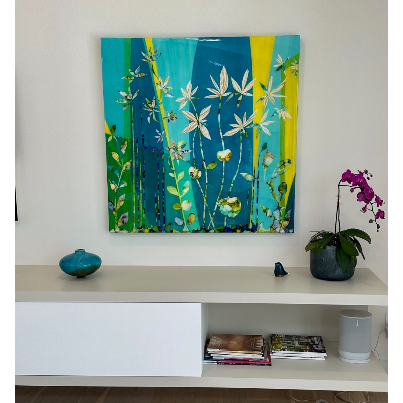 A modern white shelf with decorative items in front of a white wall. A large colorful abstract painting of botanical elements hangs on the wall. On the left side of the shelf, a blue-green round vase. On the right, a potted orchid with pink flowers i