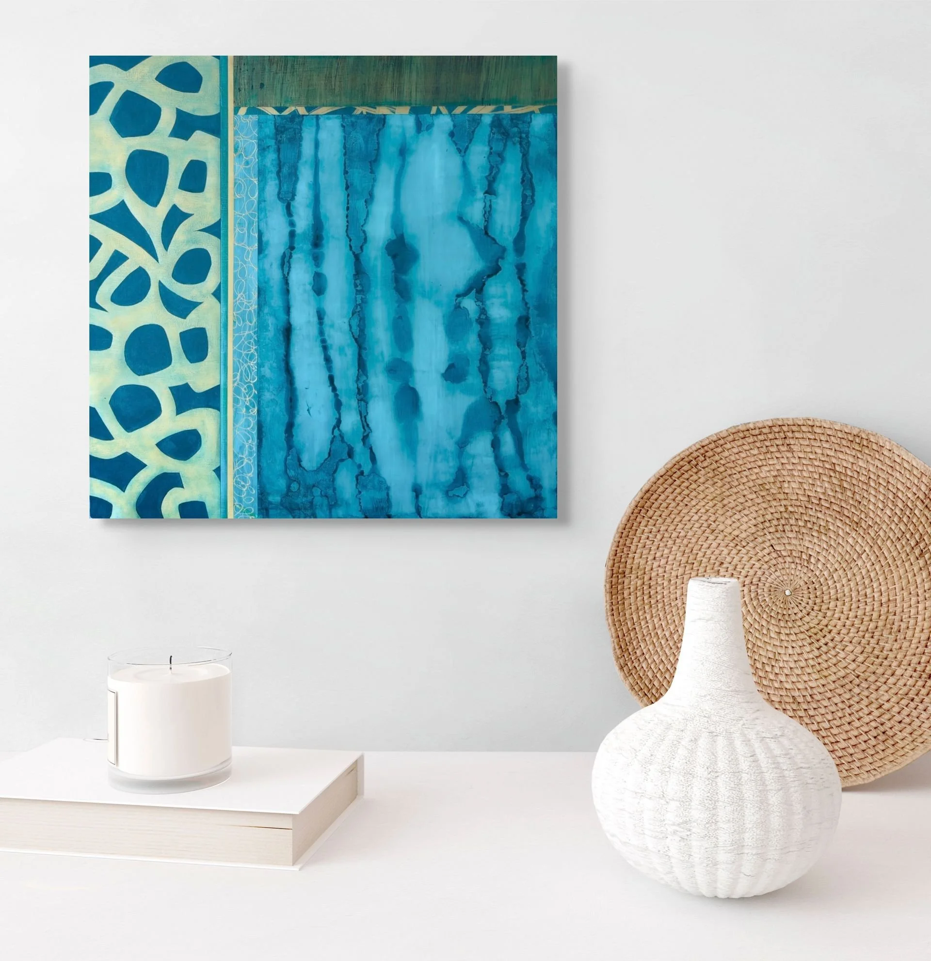 Interior scene with a white table, a white candle in a glass holder, a white textured vase, a woven round wall decor, and an abstract blue painting on a white wall.
