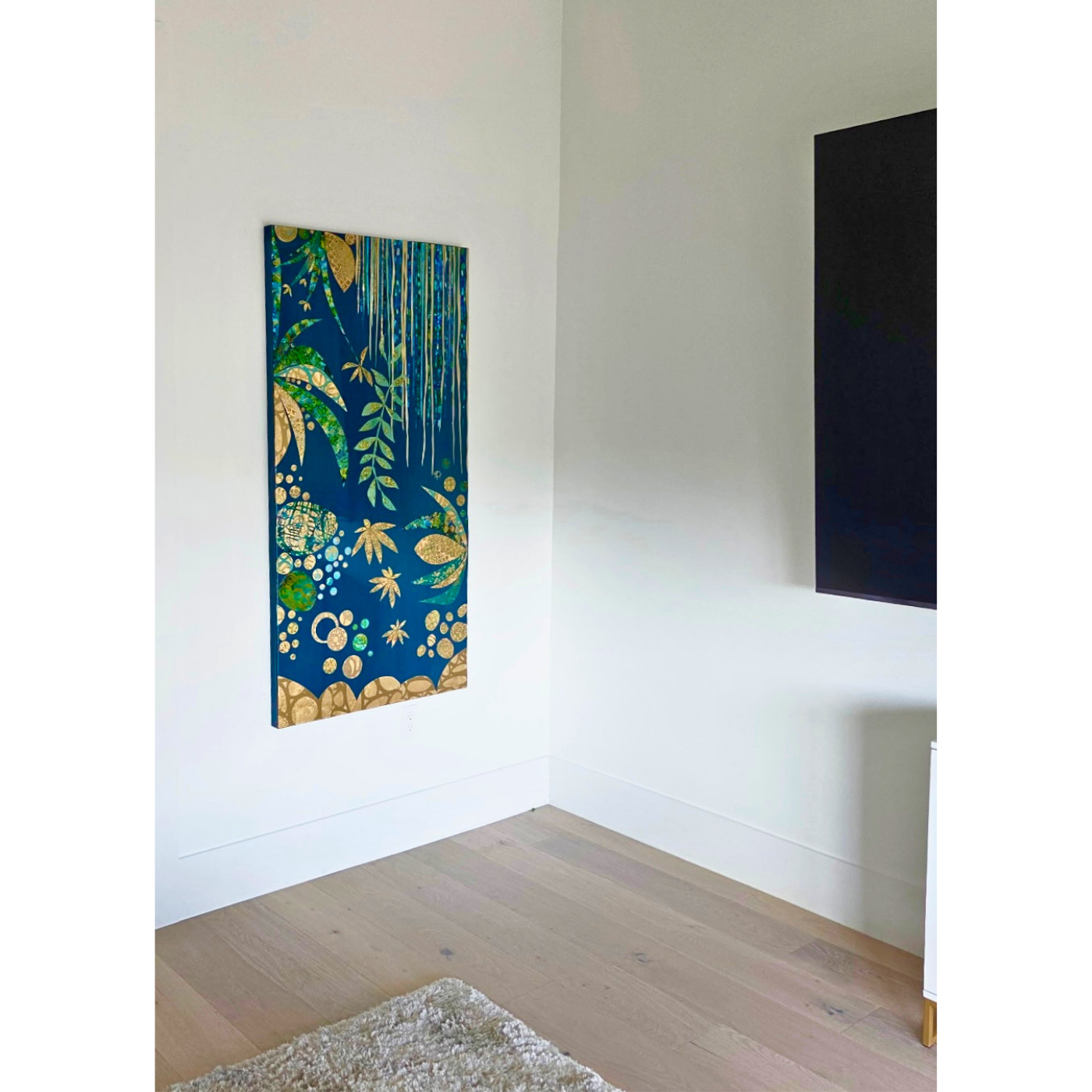 A colorful artwork with blue, green, and gold patterns of plants, leaves, and abstract shapes hanging on a white wall in a modern interior space.