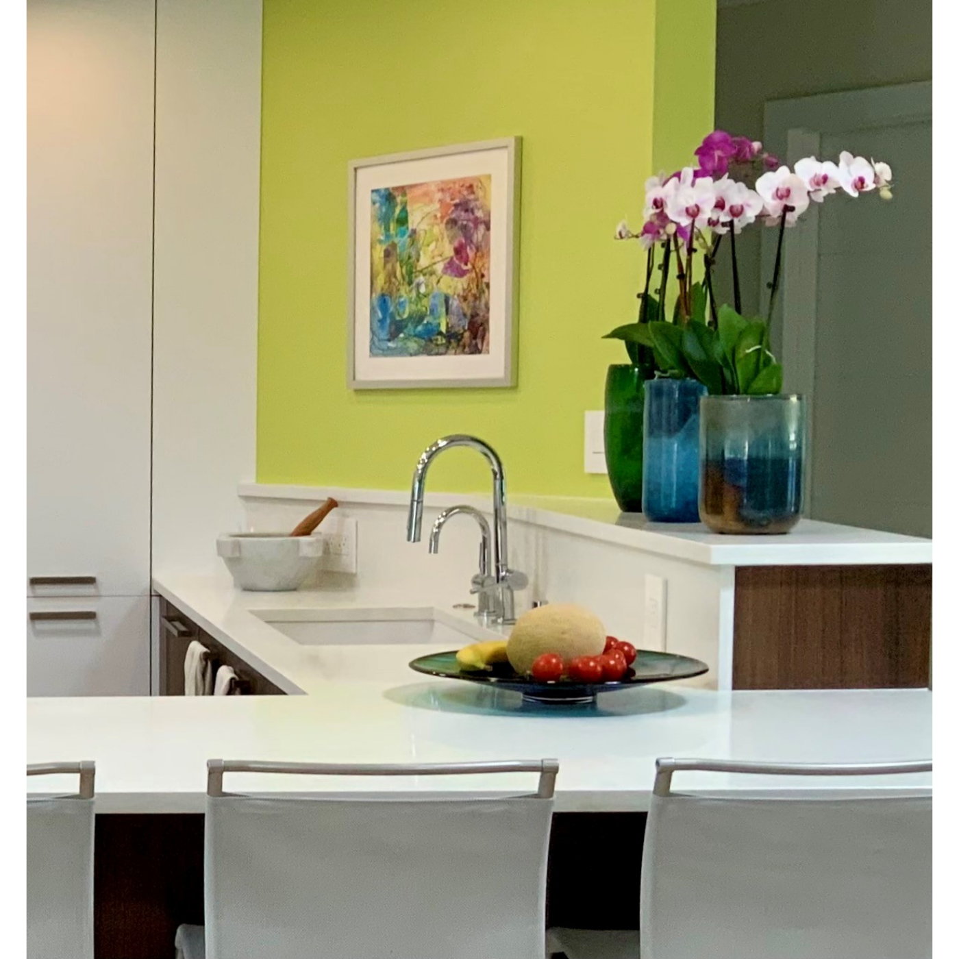 Modern kitchen with white island counter, colorful vases with orchids, a abstract art piece on a lime green wall, and a bowl of fruits including melon, banana, and cherry tomatoes.