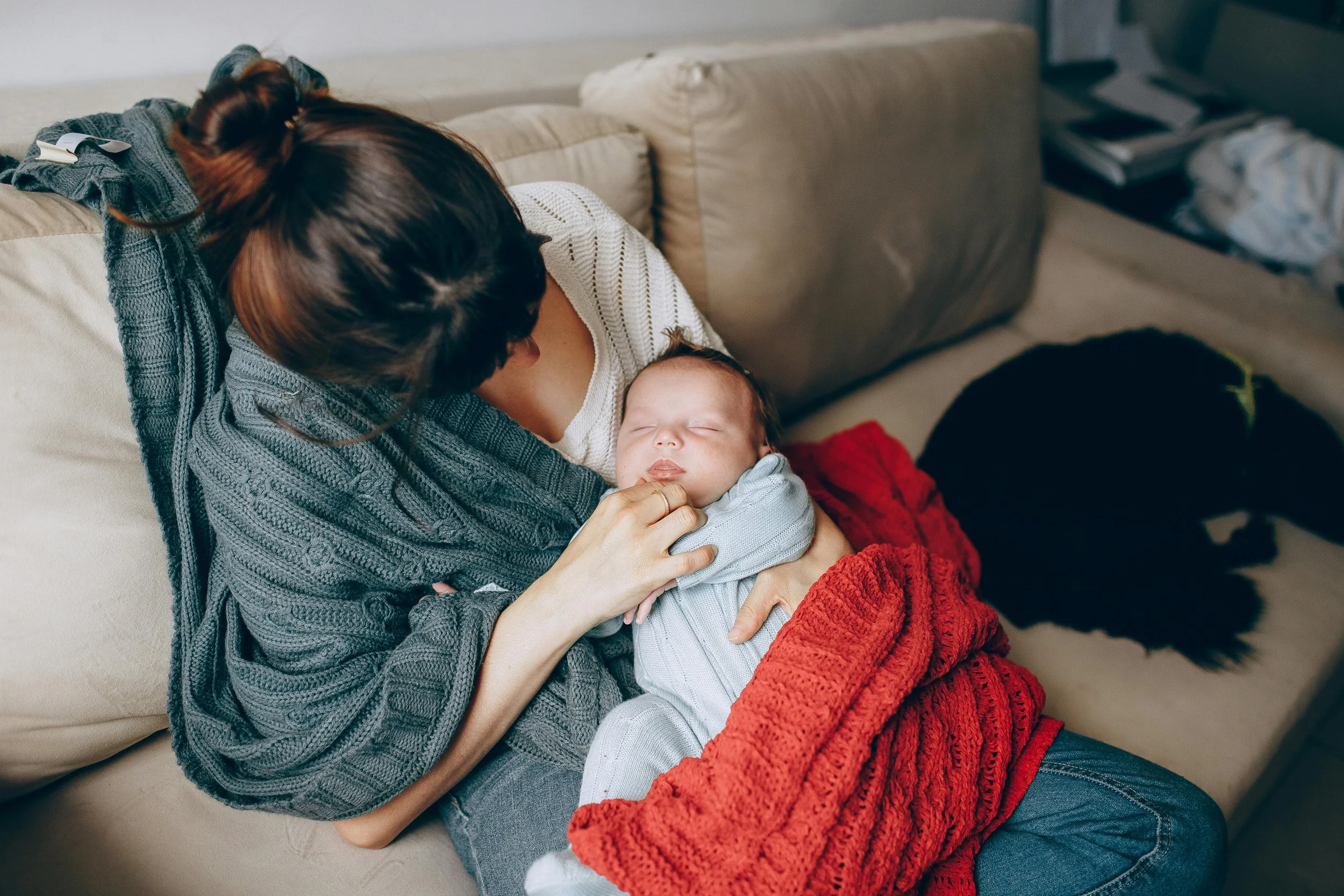 Woman lying on a couch with a baby, breastfeeding, wearing a white sweater, with a red blanket, and a gray sweater draped over her, in a cozy living room.