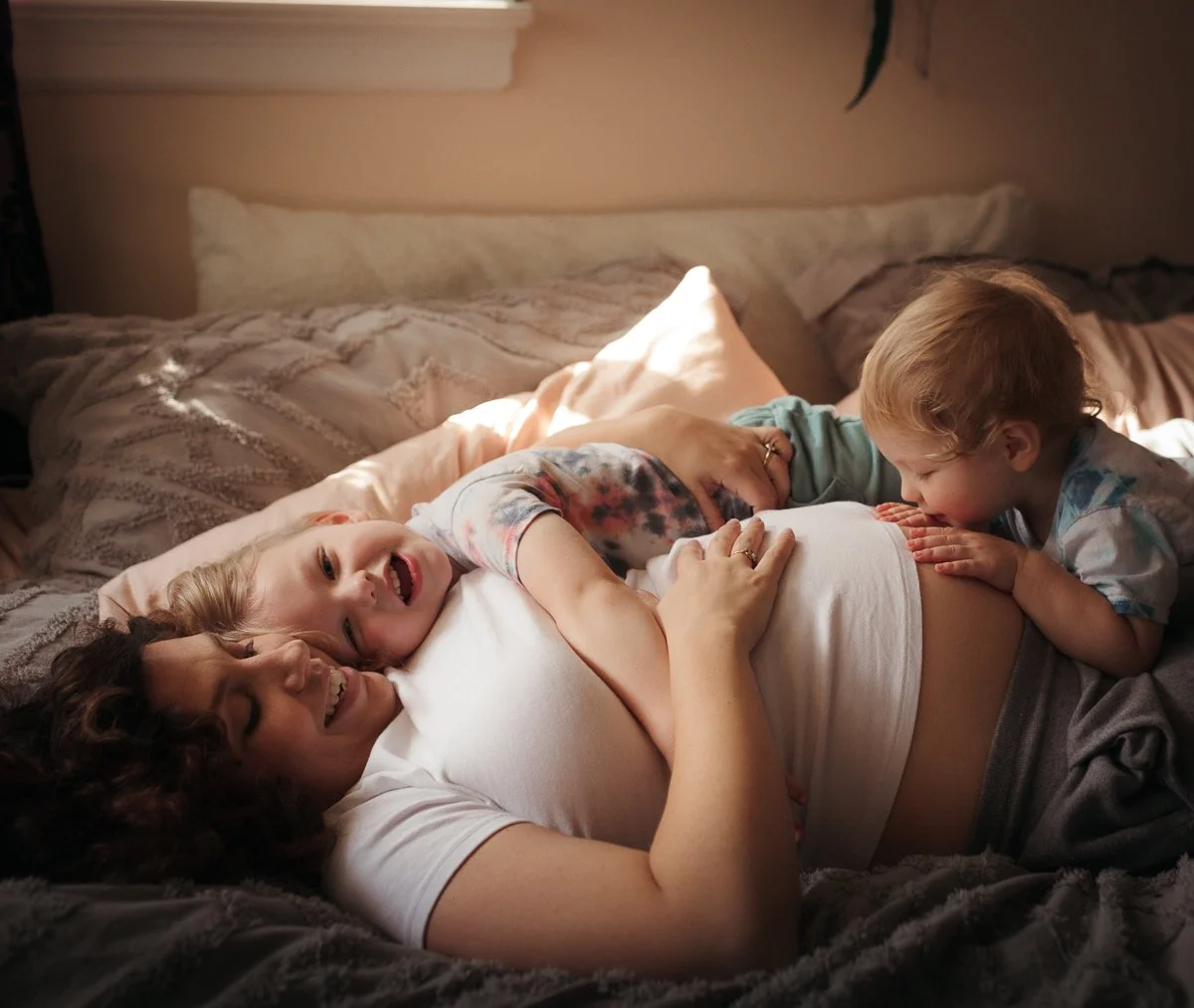 A woman lying on a bed with two young children, one boy and one girl, playfully touching her stomach and smiling in a cozy bedroom.