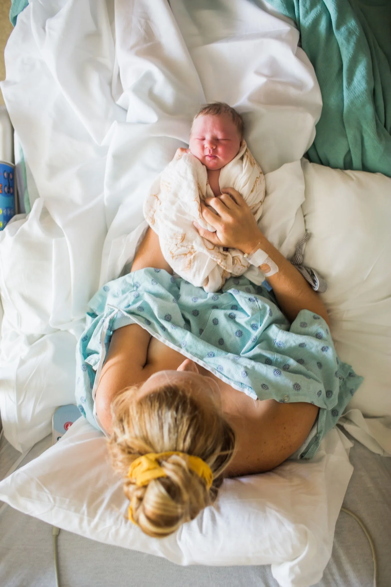 A newborn baby wrapped in a blanket lies in a hospital bed, holding a hand ultrasound monitor with a caregiver supporting them. The baby has closed eyes and a slightly furrowed brow, with a person with blonde braided hair leaning over nearby.