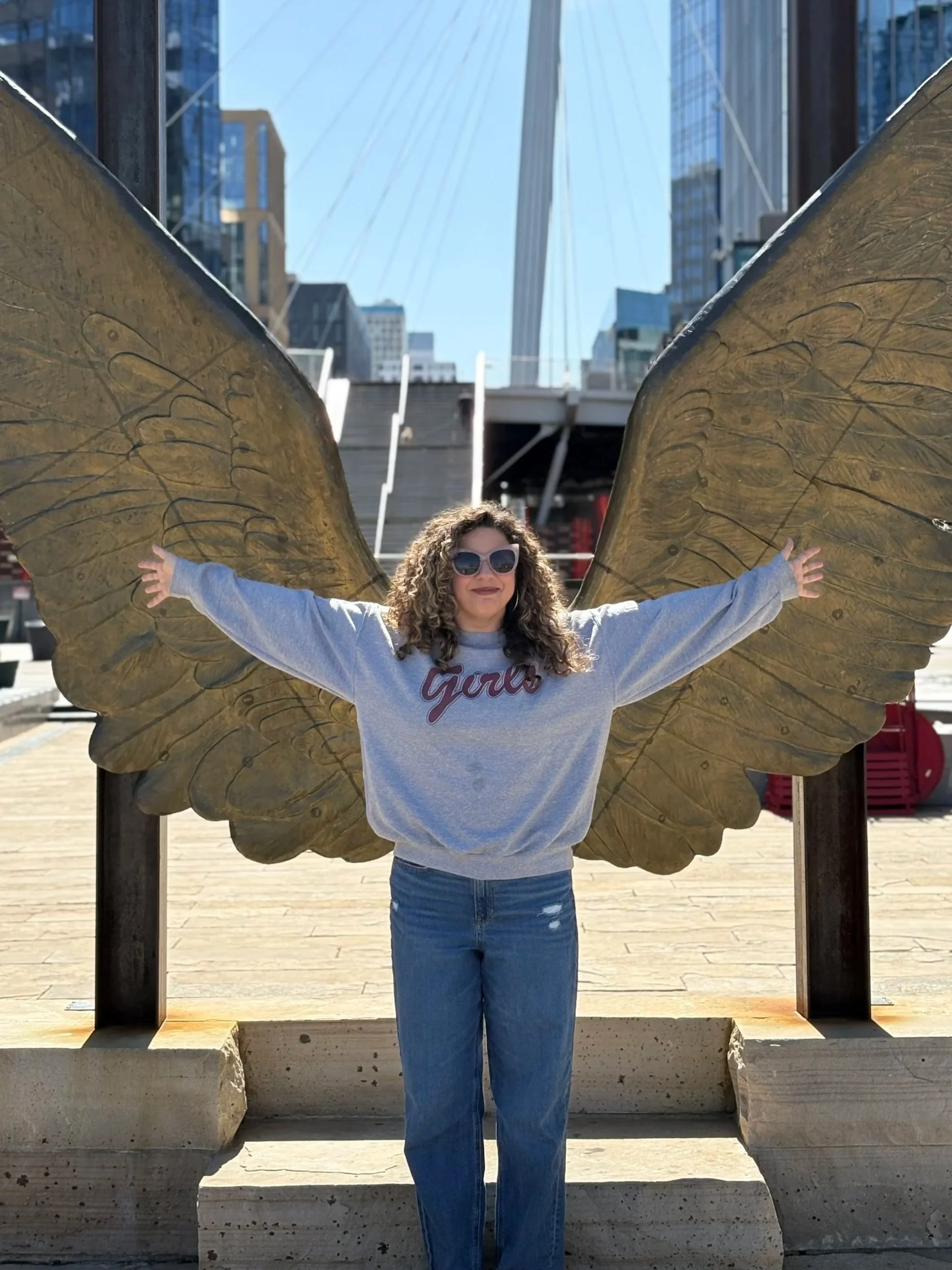 A woman with curly hair wearing sunglasses and a gray sweatshirt with a word on it, standing in front of a large sculpture of butterfly wings outdoors in an urban area with tall buildings and a bridge in the background.