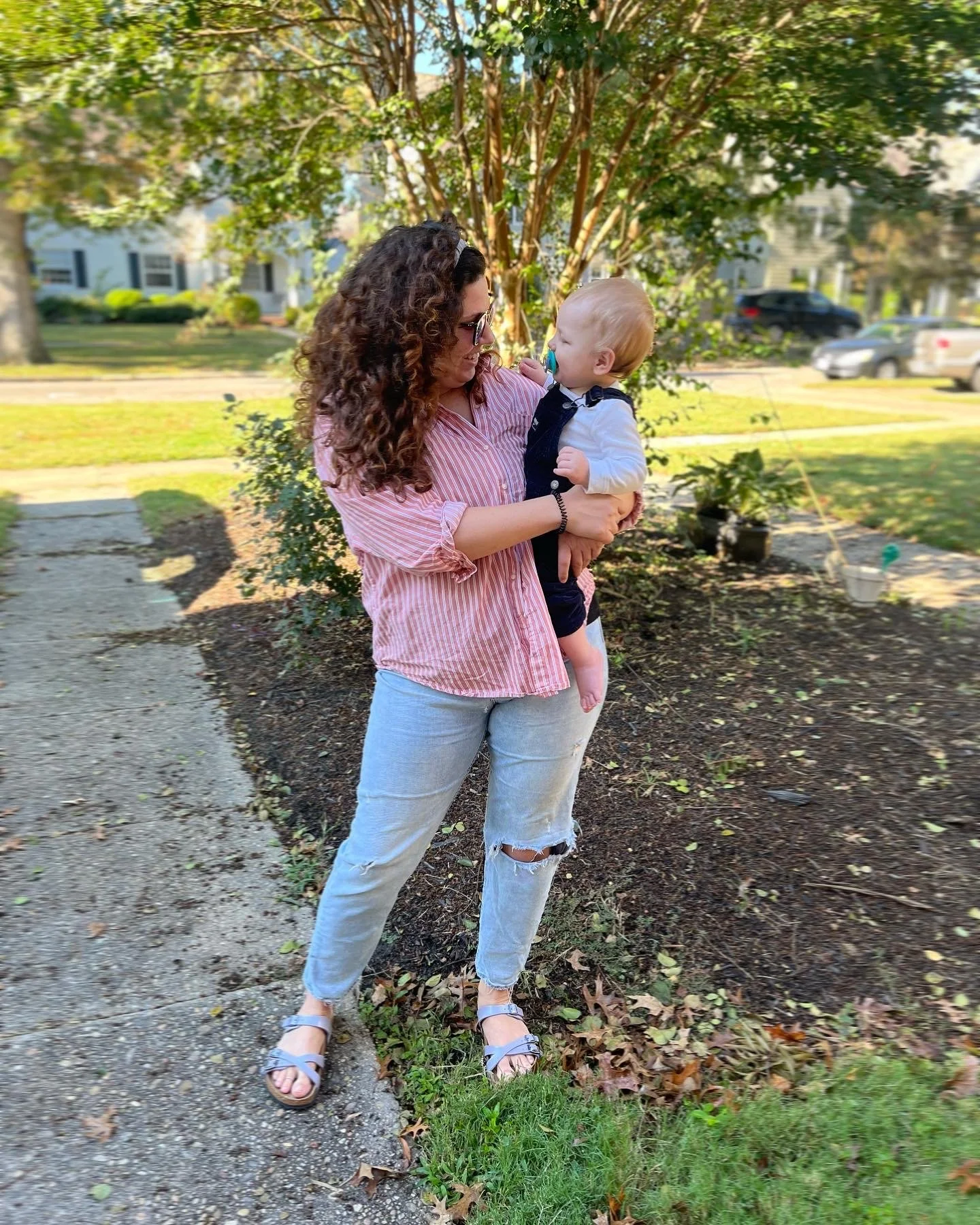 A woman with curly brown hair, wearing a pink and white striped shirt, light blue ripped jeans, and sandals, holding a young child with blond hair, a pacifier, white shirt, dark overalls, in a park with trees, grass, and parked cars in the background.