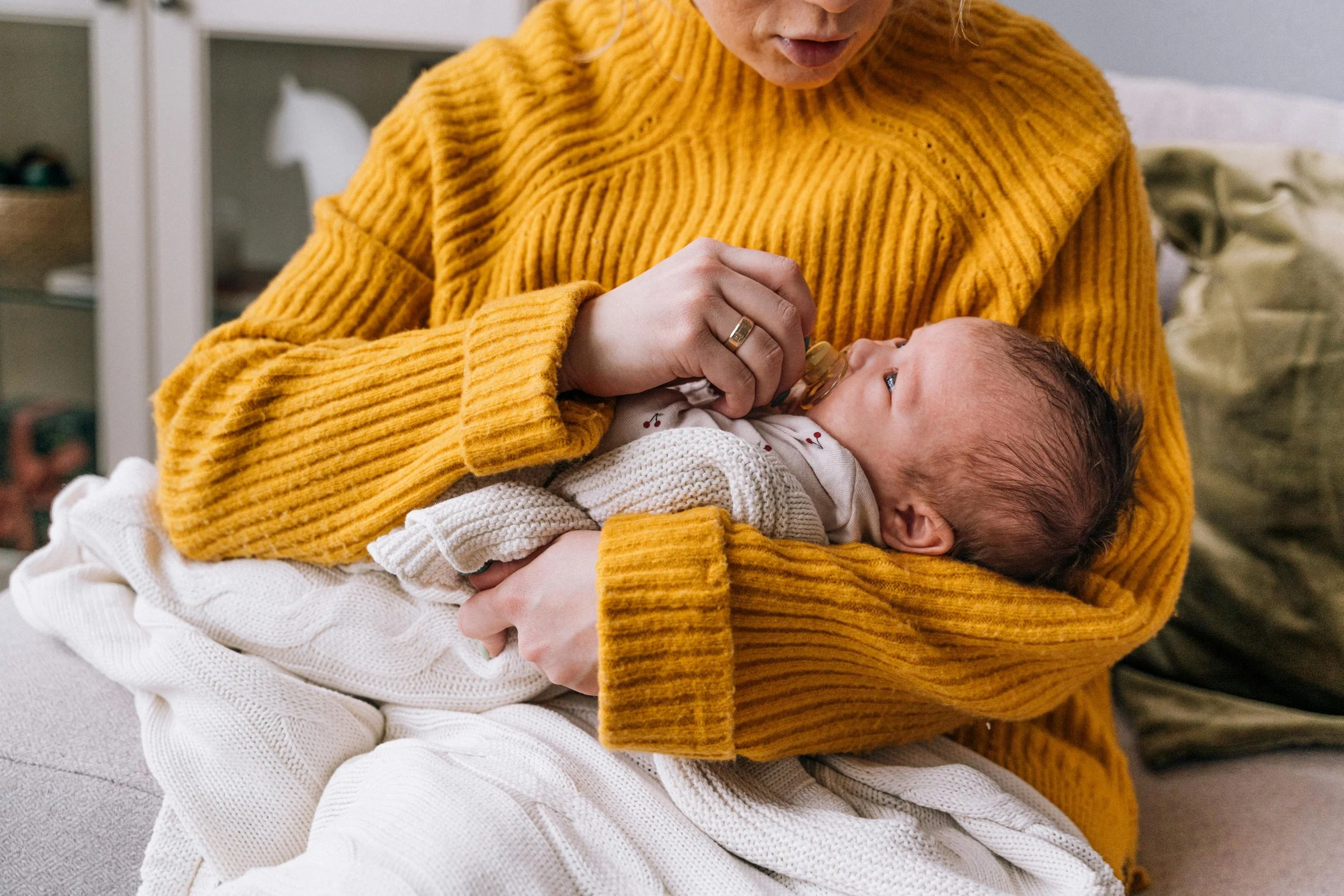 A woman in a yellow knitted sweater holding a baby in white clothing, feeding the baby with a bottle in a cozy indoor setting. birth-doula-newport-news-virginia-kinfolk-birth-co