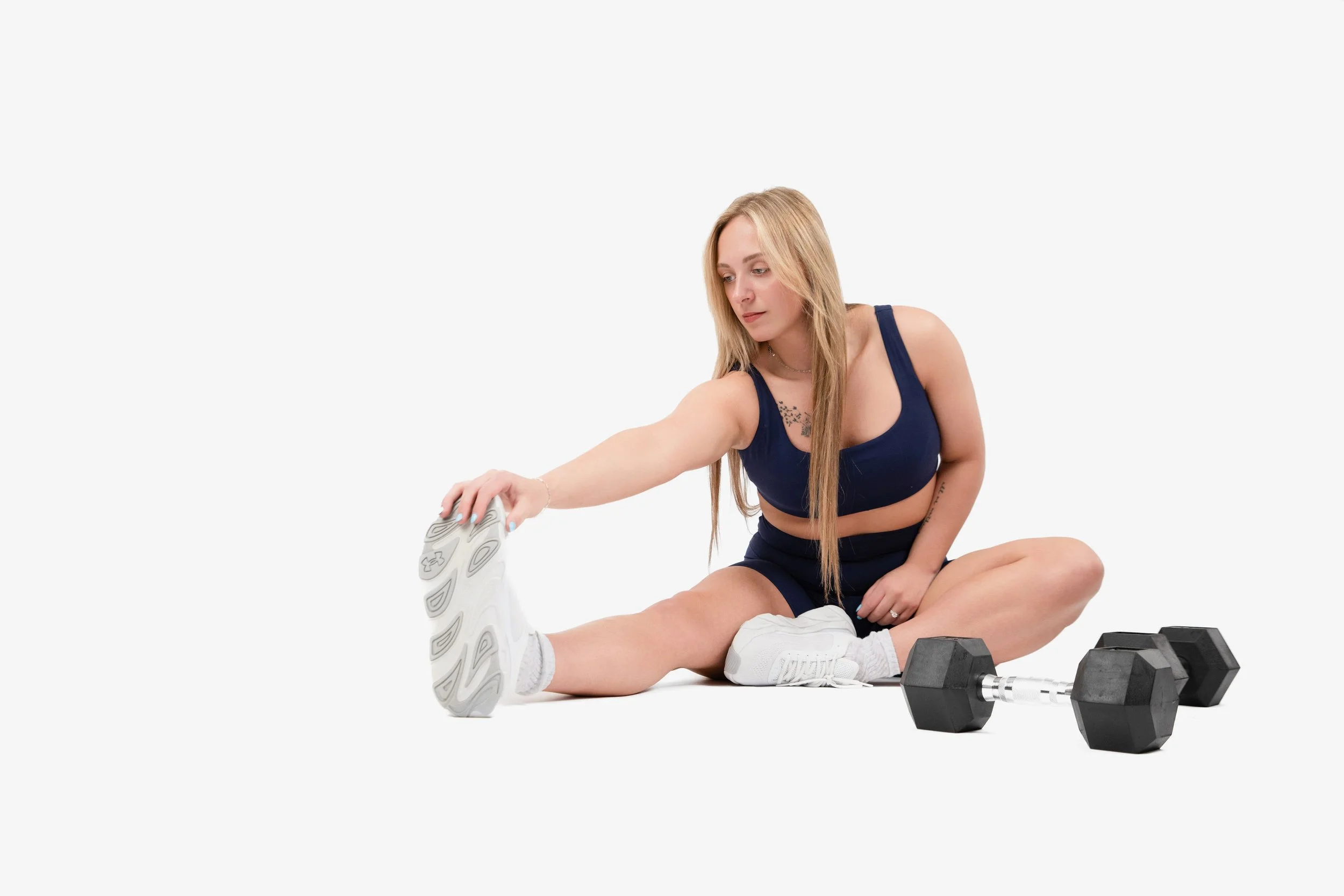 A woman with long blonde hair, wearing a blue sports bra and shorts, sitting on the floor and stretching her leg, with dumbbells nearby, against a plain white background.
