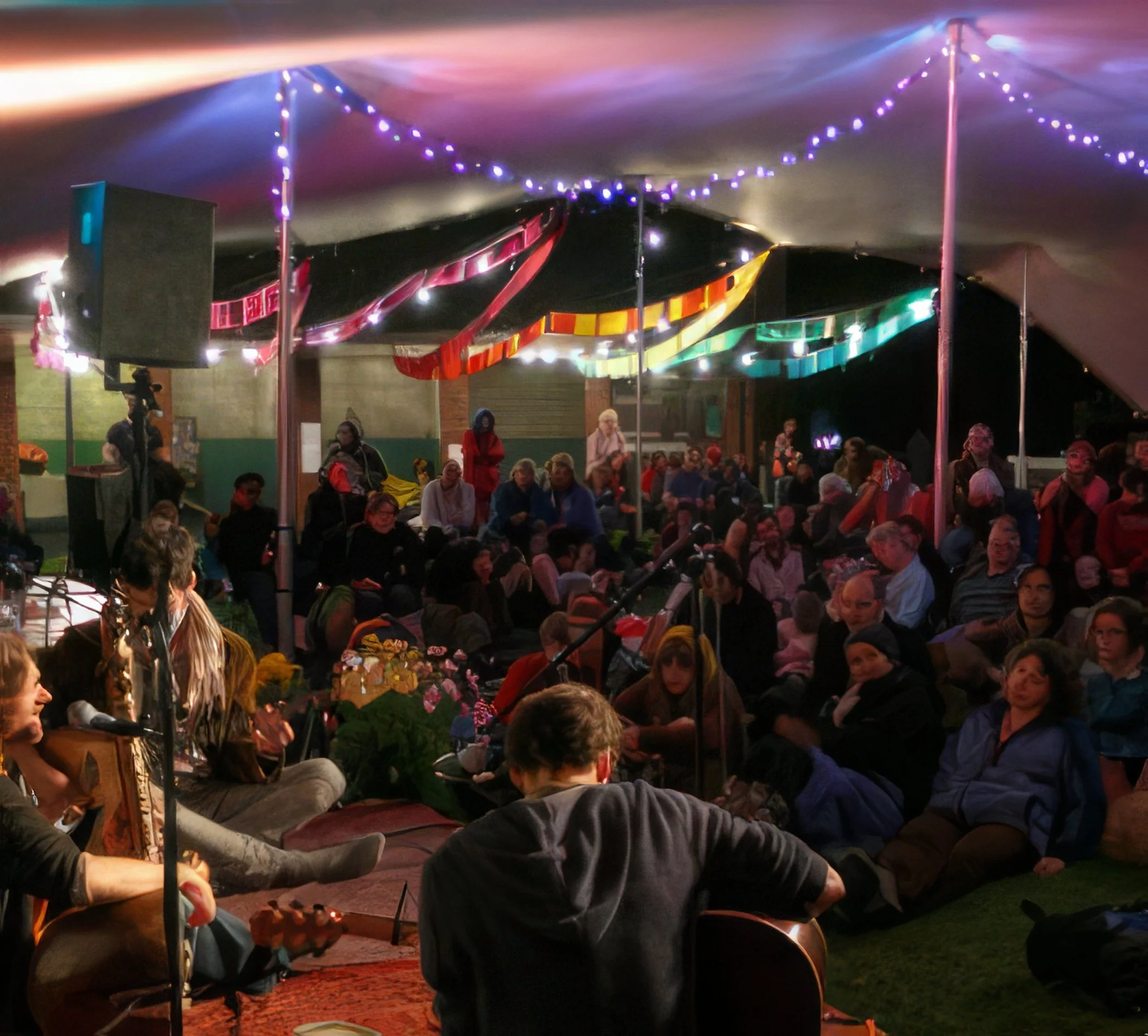 A crowd gathered under a large tent decorated with colorful lights at a nighttime outdoor event, with some people sitting on the ground and others standing, while musicians perform near the front.