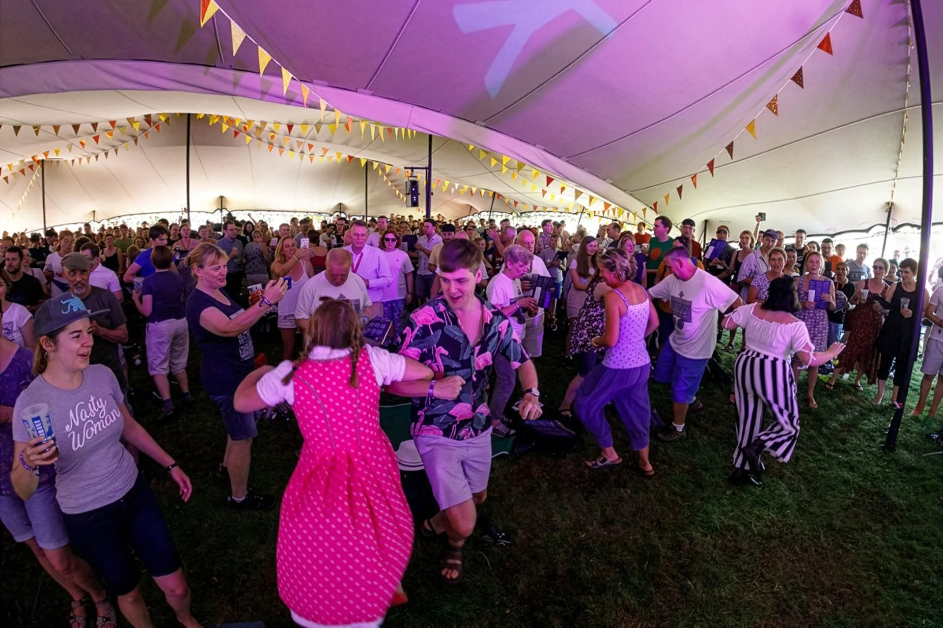 People dancing and socialising under a large stretch tent at a festival or celebration, with bunting decorations hanging from the ceiling.