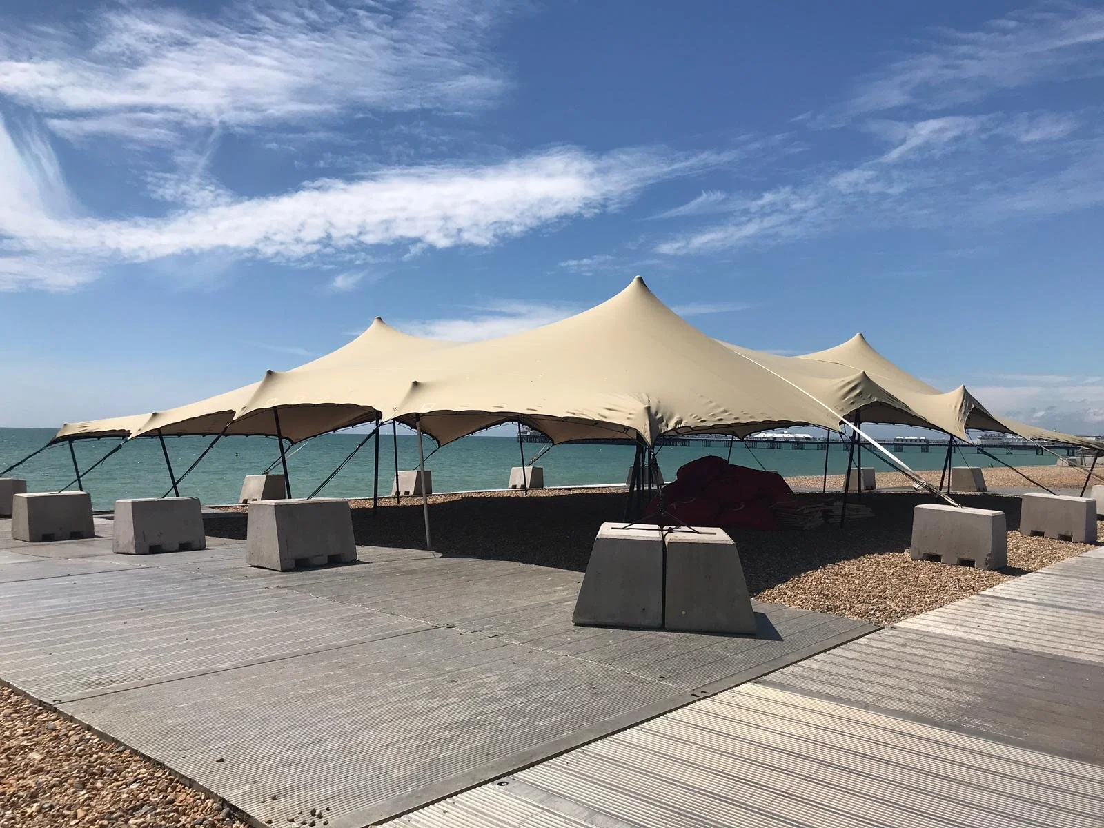 Beachside scene with large stretch tent and a wooden walkway near the water, under a partly cloudy sky.