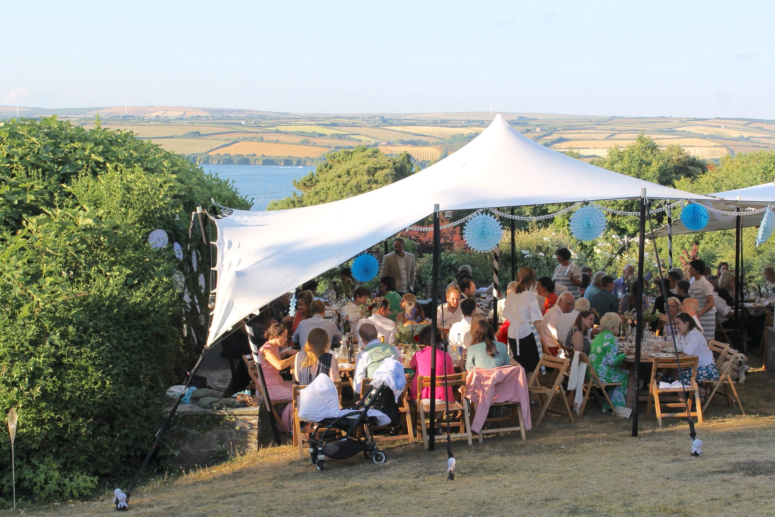 Outdoor gathering of people under a white stretch tent decorated with blue and white paper lanterns, overlooking a body of water and rolling hills.