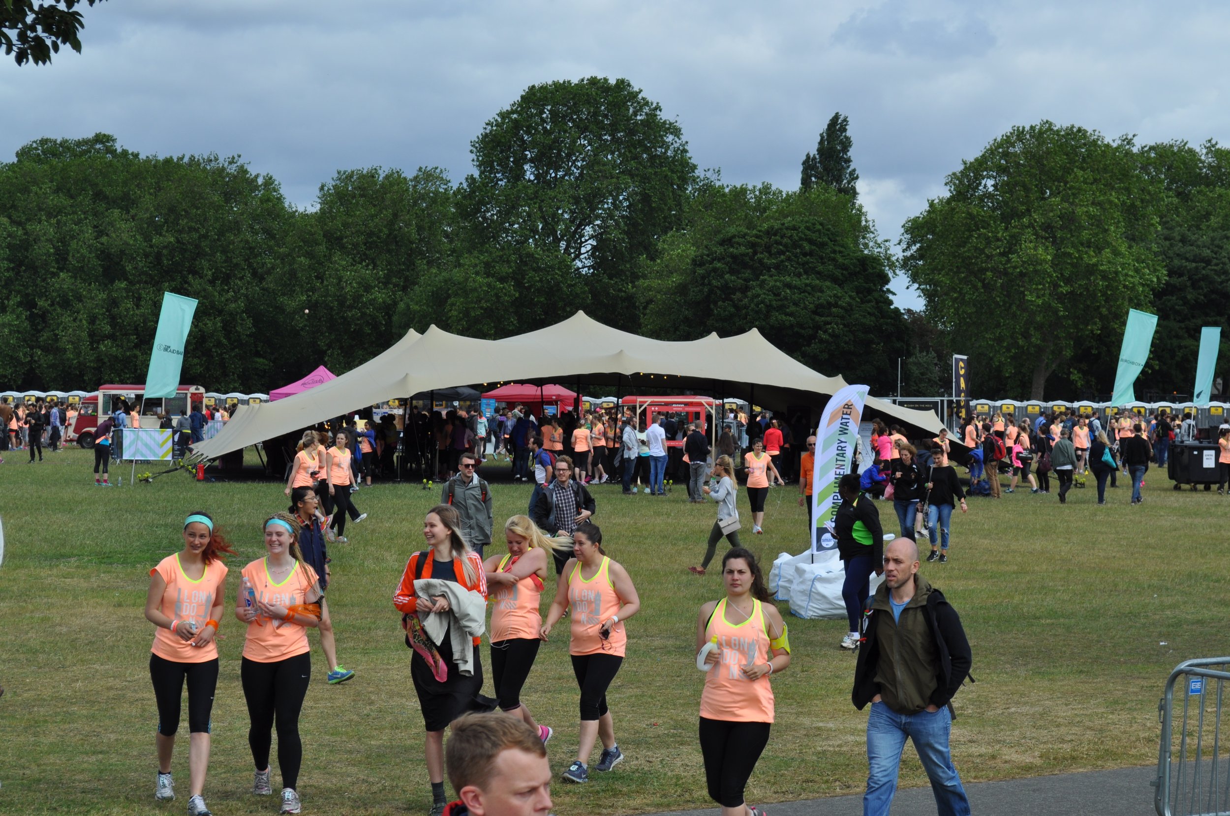 Crowd of people attending an outdoor event in a park with a large white stretch tent and green trees in the background.