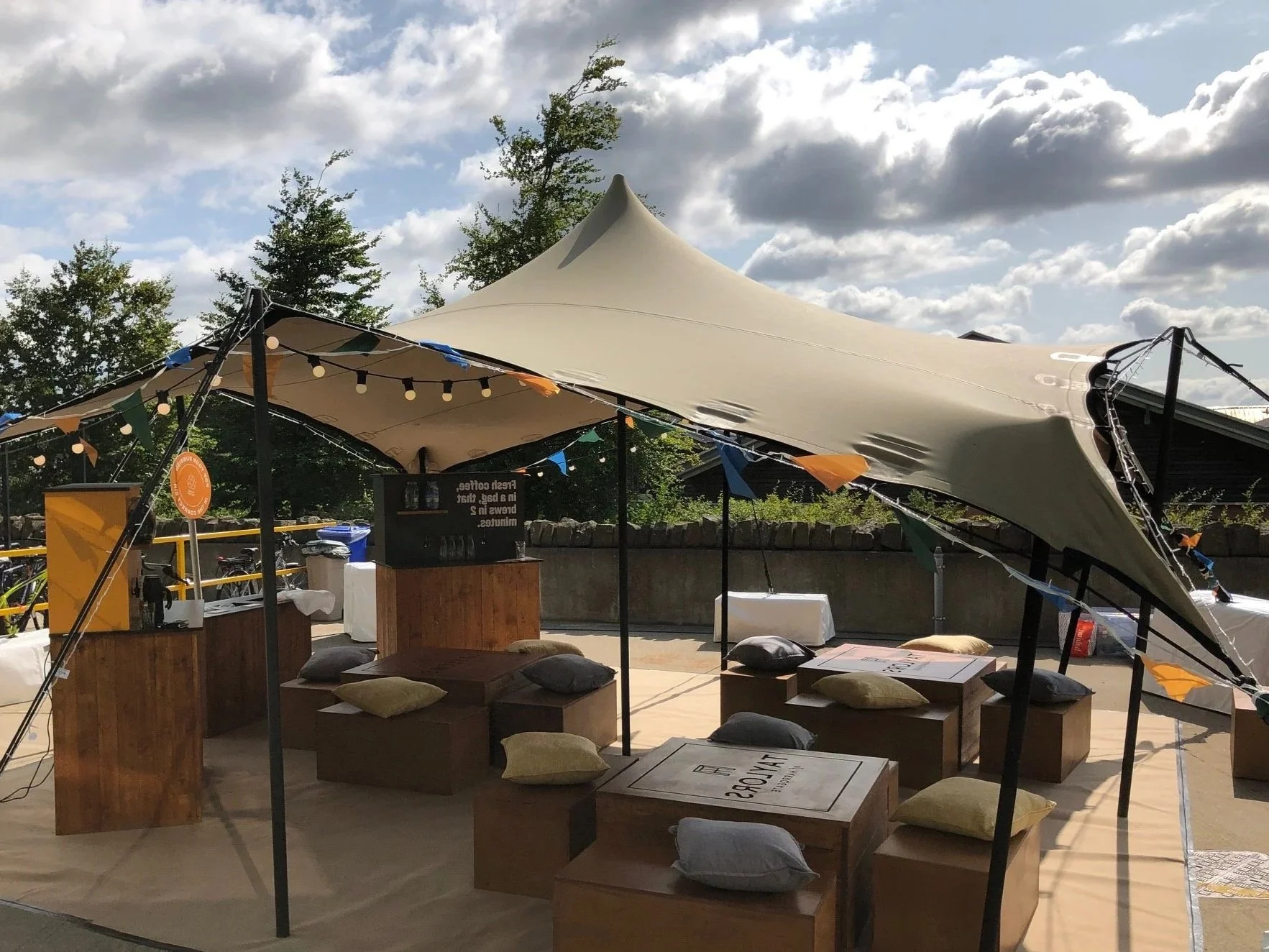 Outdoor event setup with a large beige stretch tent, string lights, wooden tables, and cushions, under partly cloudy sky.