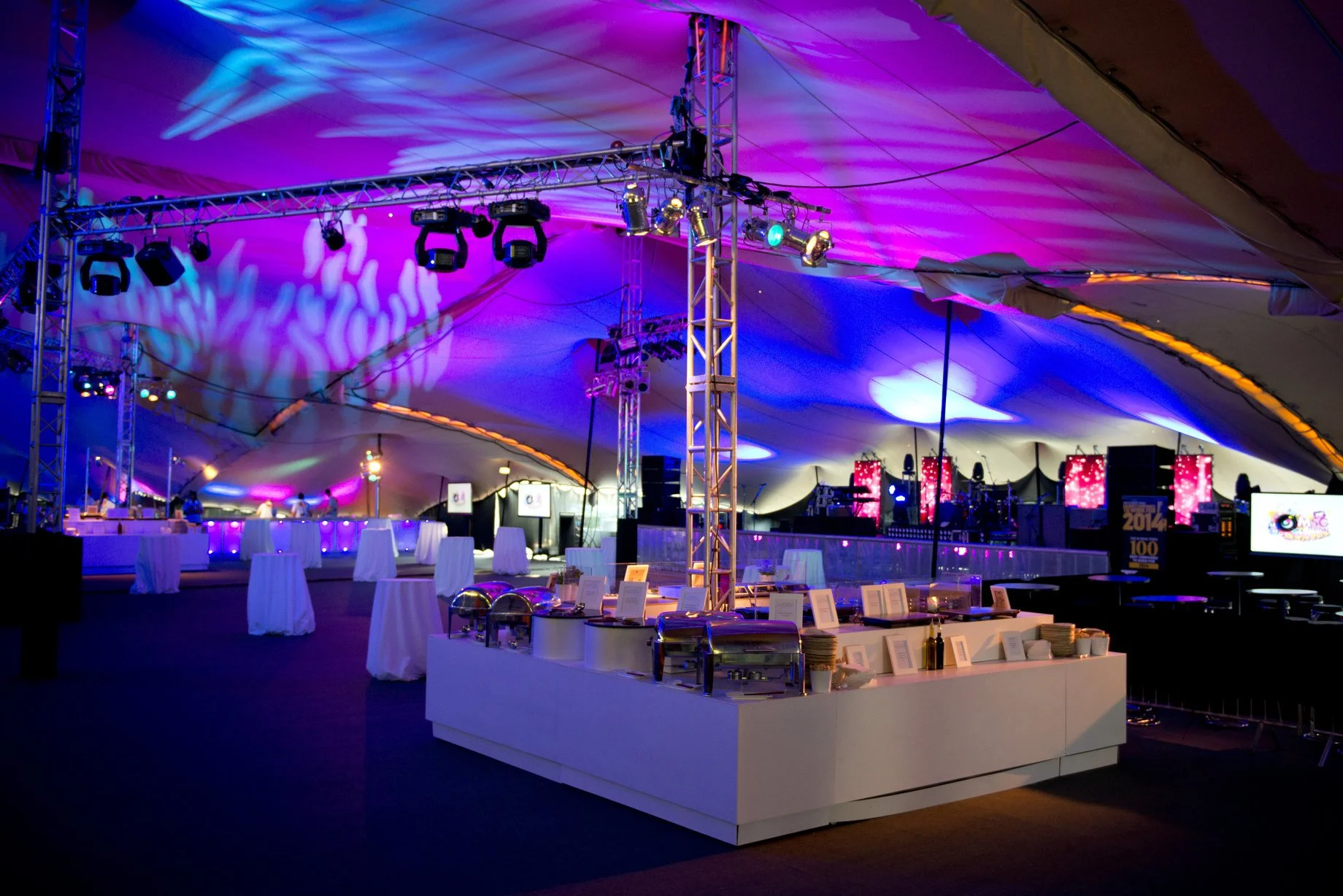 Interior of a large  stretch tent decorated with purple and blue lighting, featuring a buffet table with chafing dishes, standing tables draped with white cloths, and a stage in the background with musical equipment and screens.