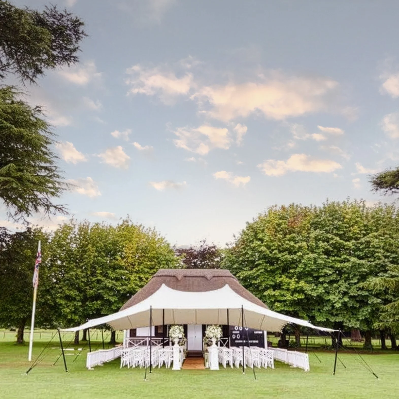 An outdoor wedding setup with a white stretch tent on a grassy field, surrounded by trees, with white chairs arranged in rows. There is a black signboard near the entrance of the tent and an American flag on a pole to the left.