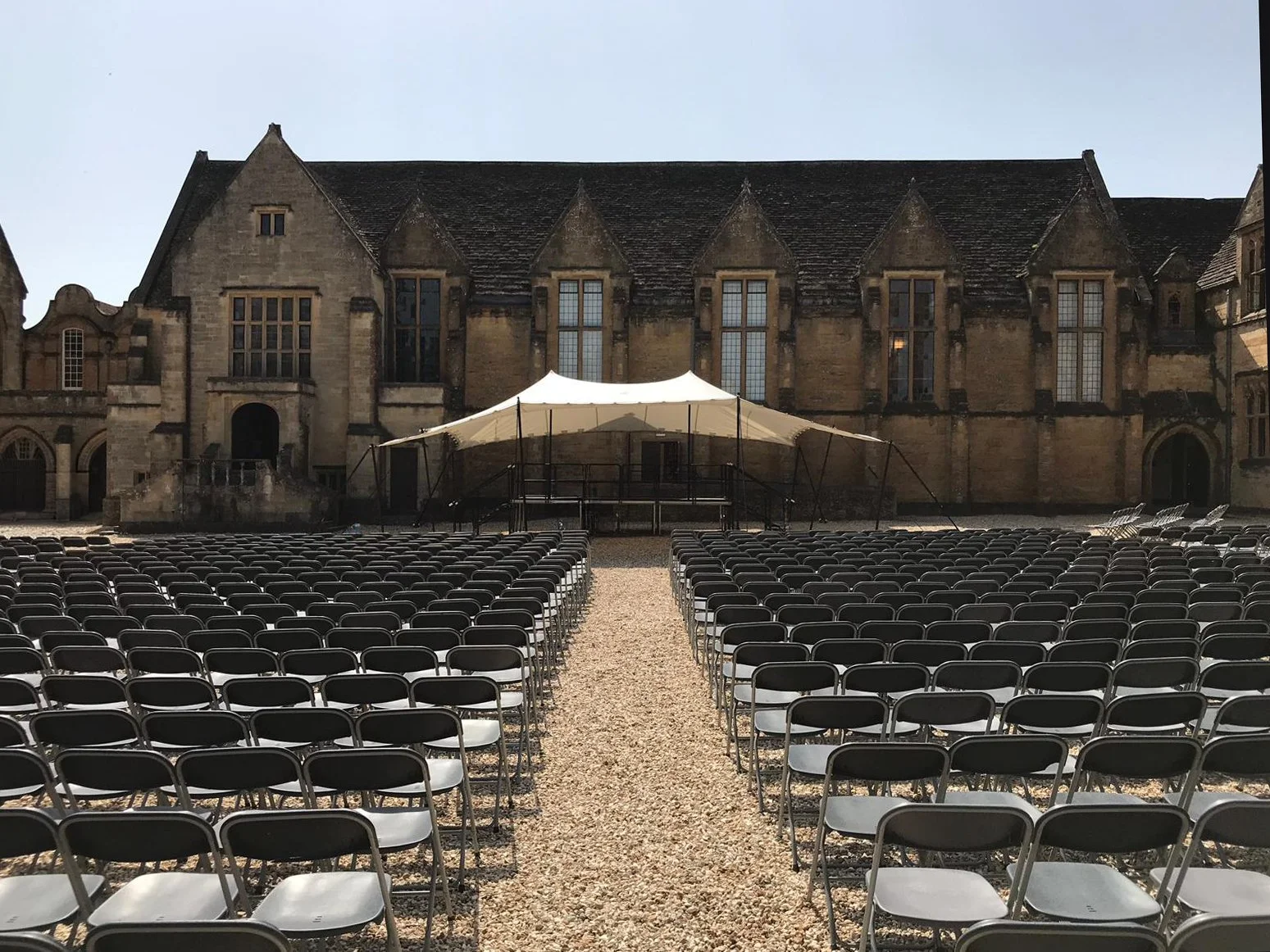 Rows of empty folding chairs facing an outdoor stage with a white canopy, in front of an old stone building.