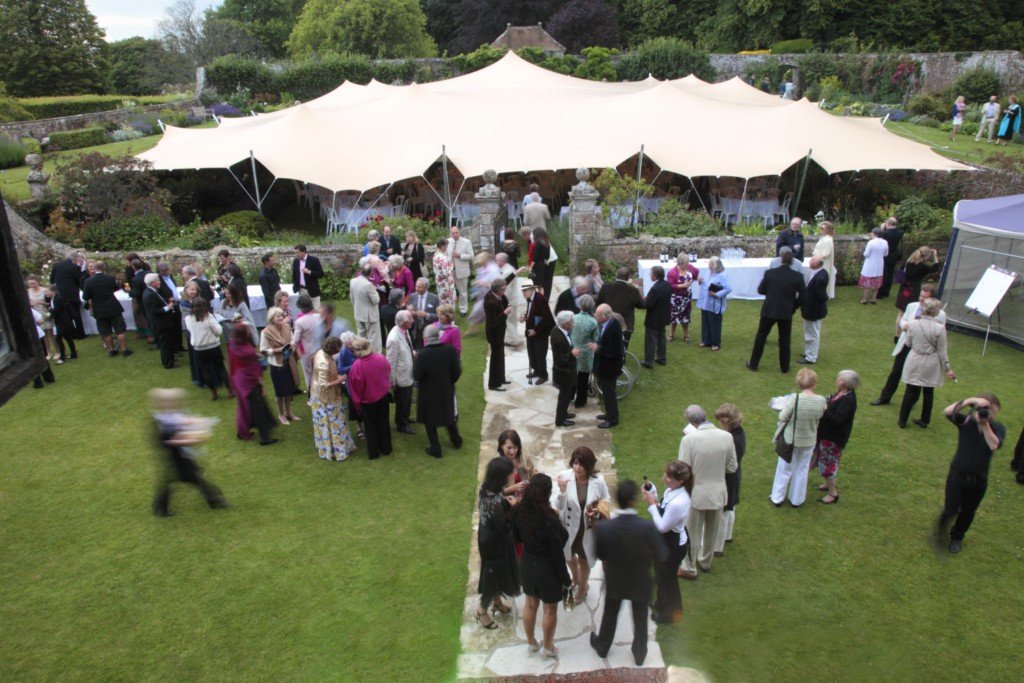 Guests gathered outdoors at a garden party or wedding reception, standing on a well-maintained lawn with a large white stretch tent in the background, surrounded by greenery and a stone wall.