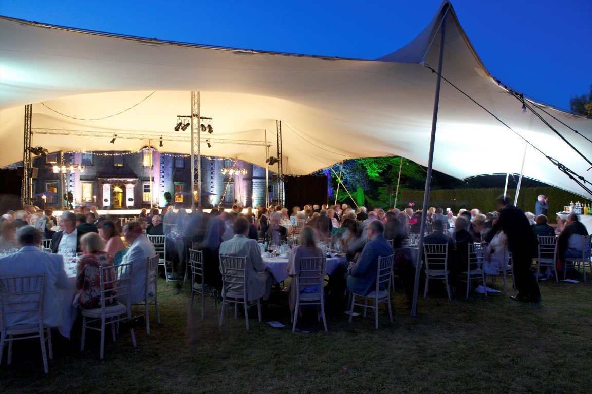 An outdoor event held under large white stretch tent with tables and chairs filled with guests, a stage with lighting in the background, and the evening sky overhead.