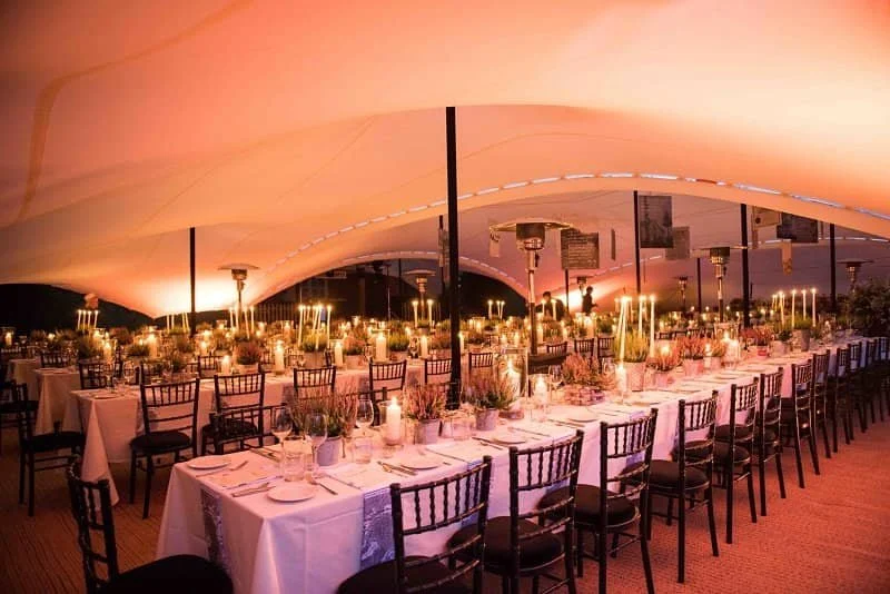 Elegant banquet table set under a large tent with pink lighting, decorated with candles, floral centerpieces, and place settings with white napkins and glassware.