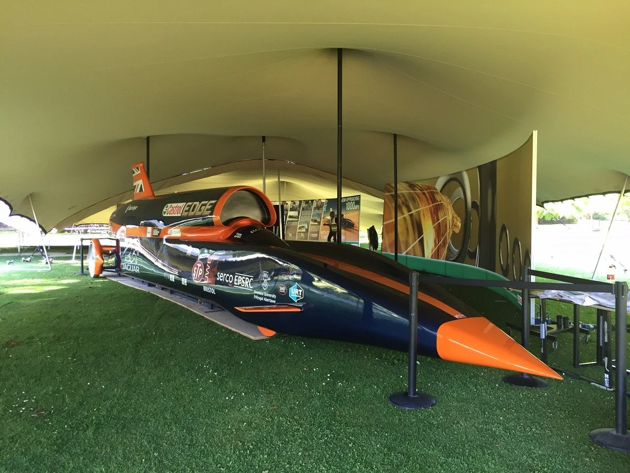 A supersonic car displayed under a large stretch tent with informational posters behind it. The car is black with orange accents and sponsor logos.