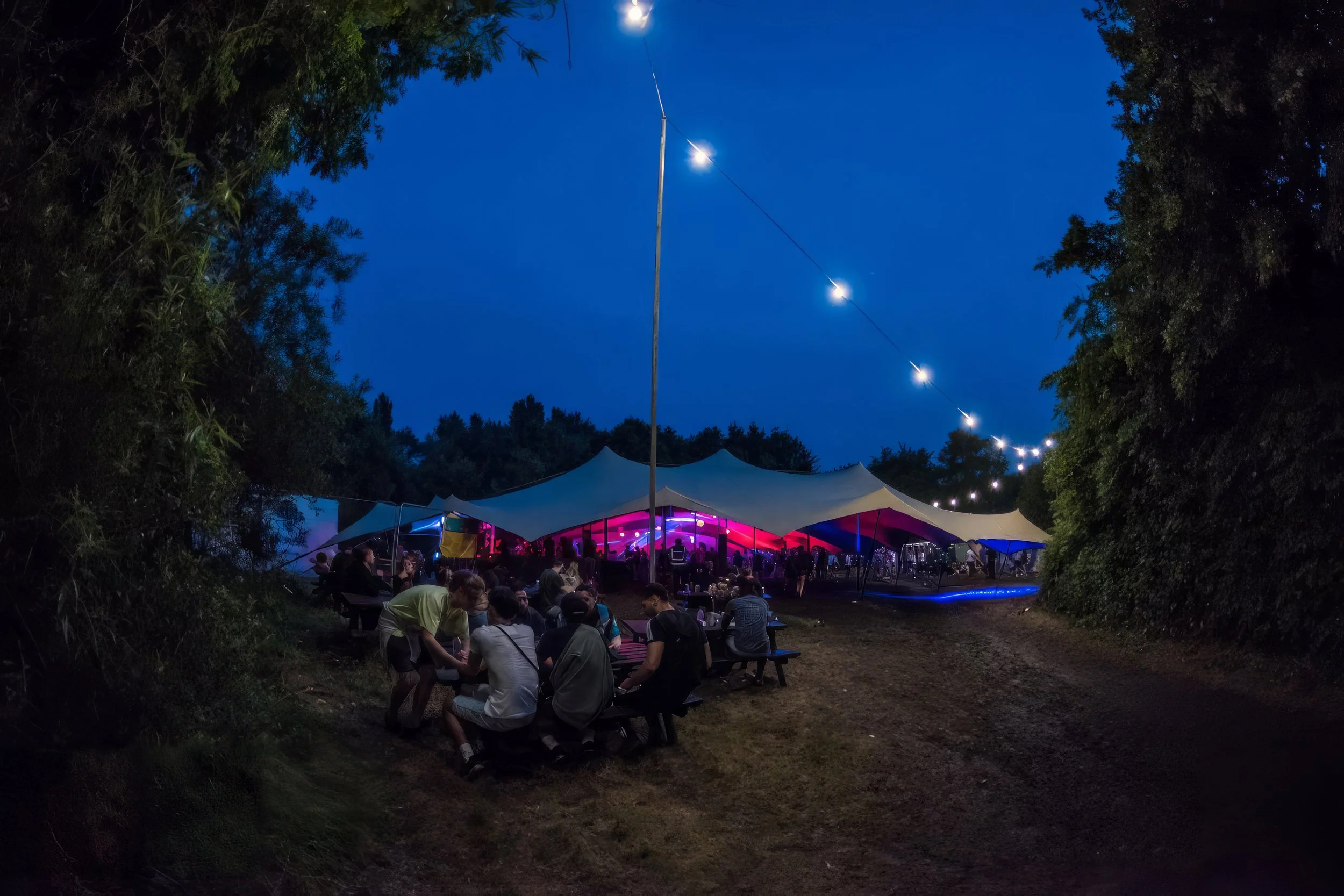An outdoor evening event with a large tent illuminated by colorful pink and purple lights, surrounded by trees and string lights overhead. People are sitting at tables and enjoying the gathering.