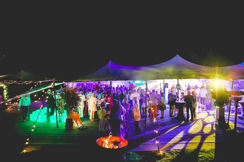 Nighttime outdoor party under a large tent with colorful lighting, people dancing and socializing.