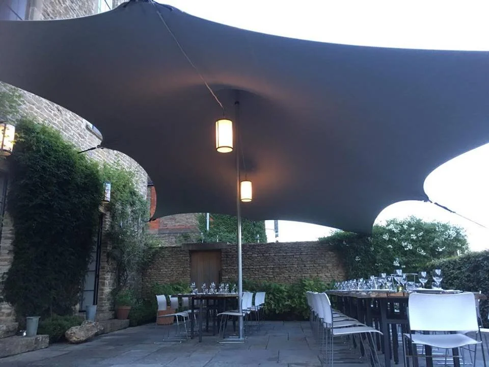 Outdoor dining area under a stretch tent, tables set with wine glasses, and chairs arranged for a gathering, surrounded by stone walls and greenery.