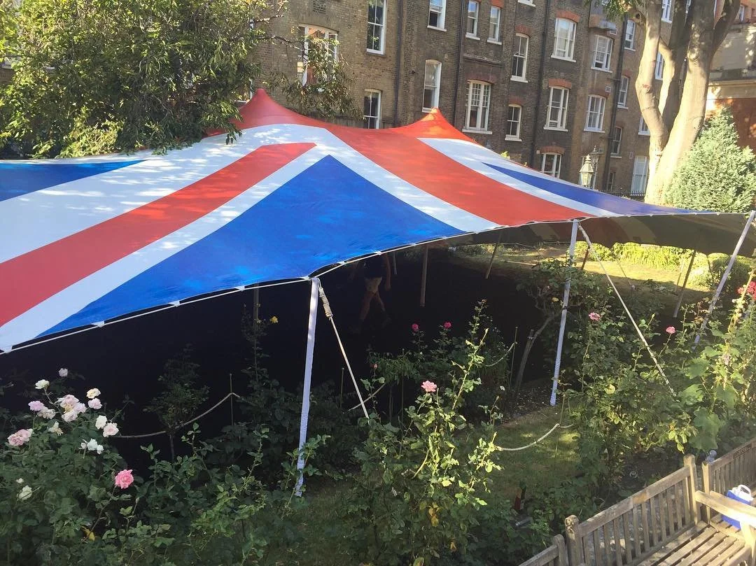 A large tent with a Union Jack design set up in a garden with rose bushes and a wooden bench.