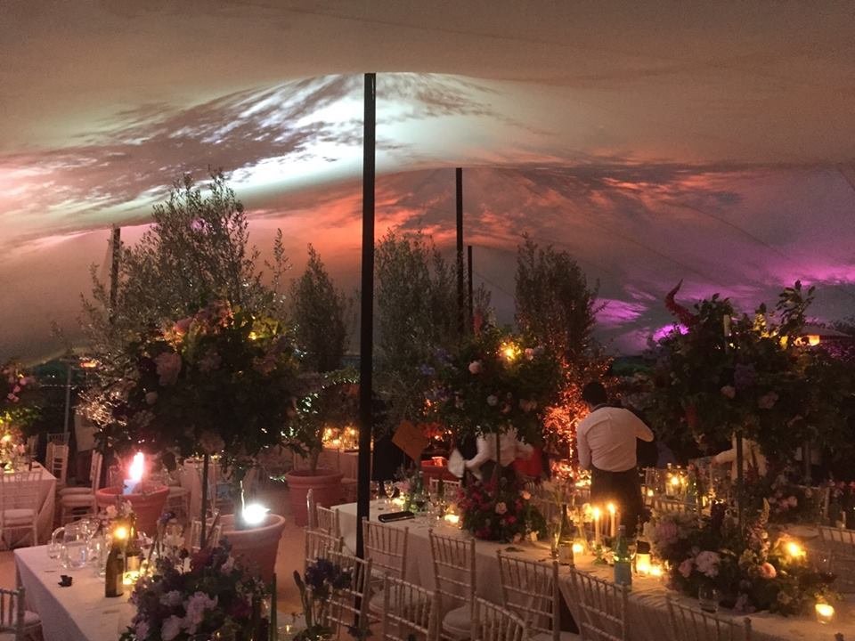 Indoor event space decorated with floral arrangements and candles, under a large stretch tent with a colourful, cloudy sky projected on the ceiling.