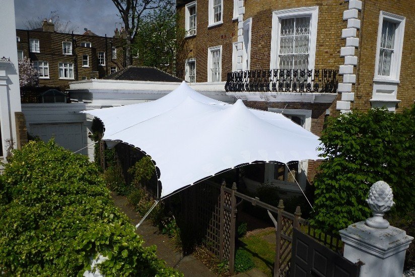 A white stretch tent tent set up in a garden area surrounded by green bushes and a black fence, with brick houses visible in the background.